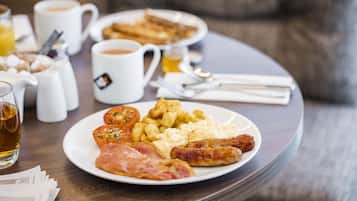 Una mesa de desayuno con huevos, salchichas, jamón y papas, junto con tazas de café y un vaso de jugo de naranja.