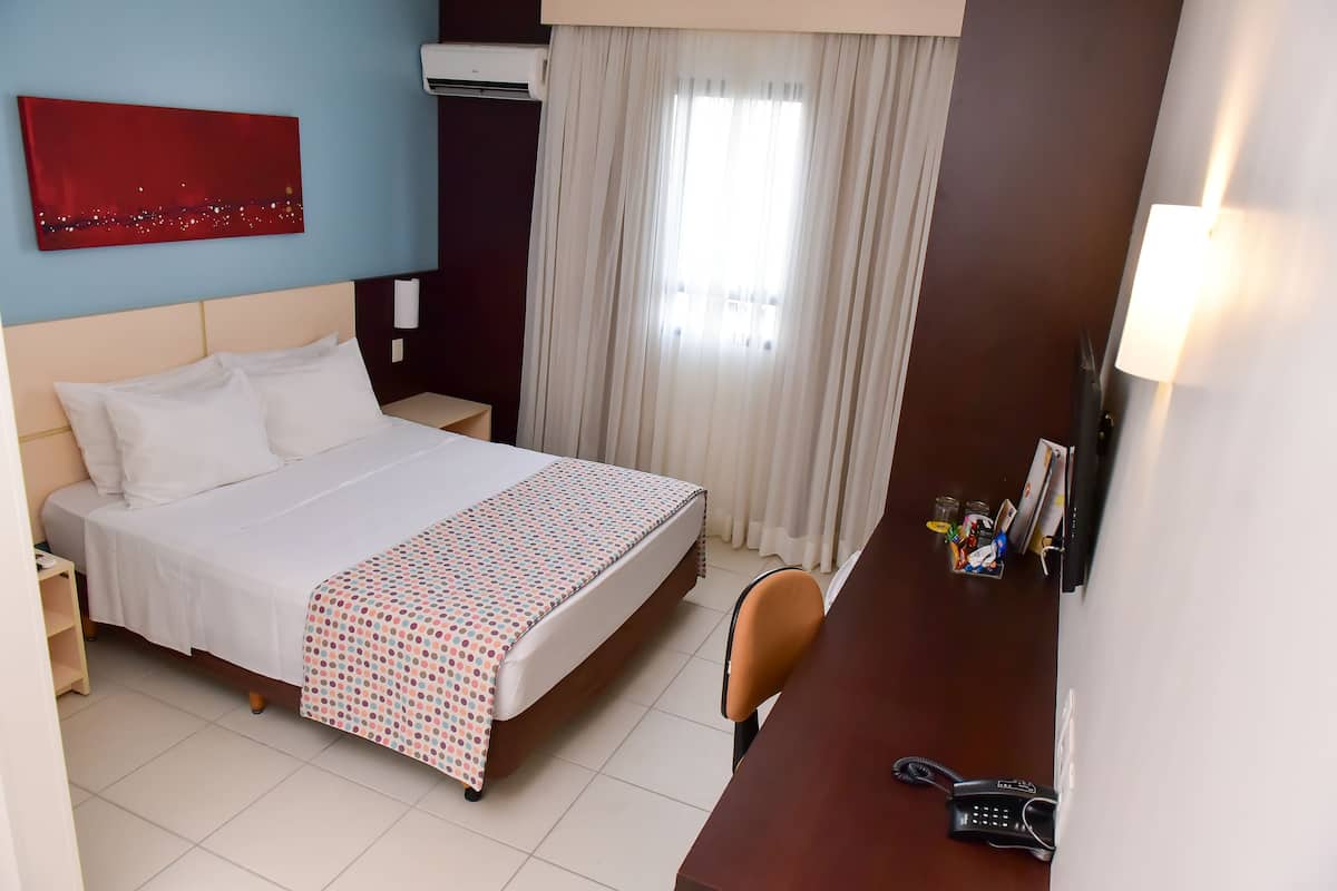 A hotel room with a bed, a desk with a phone, a chair, a nightstand, a window with curtains, and a red artwork on the wall.
