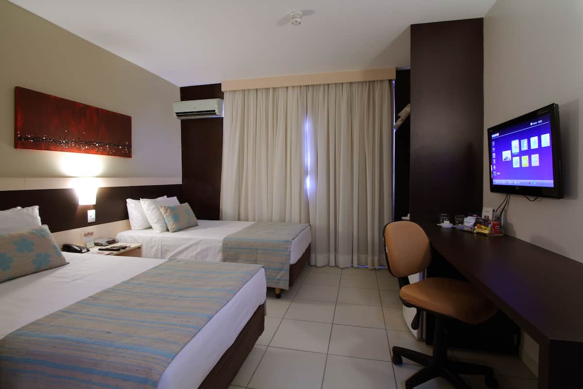 A hotel room with two beds, a desk, a chair, a TV, and an air conditioning unit.