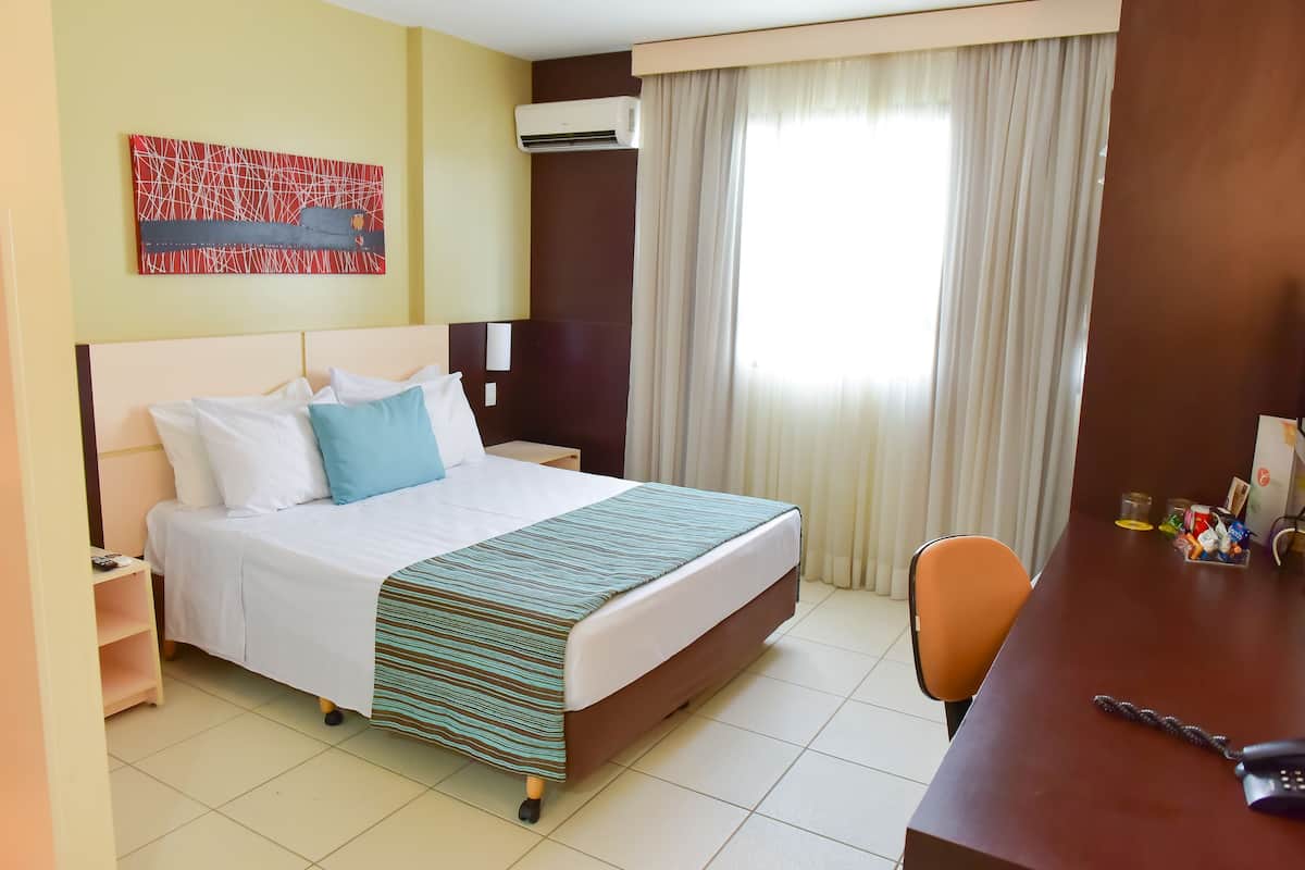 A hotel room with a bed, a desk, a chair, a nightstand, an air conditioner, a window with curtains, and a wall-mounted artwork.