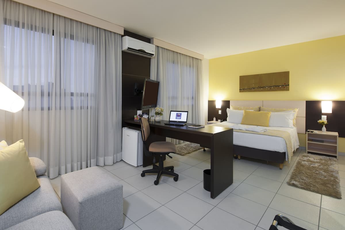 A hotel room with a bed, a desk with a computer, a chair, a sofa, and a lamp.