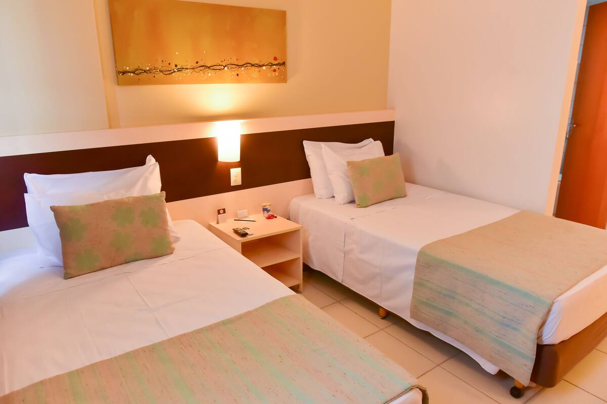 A hotel room with two single beds, a nightstand, a wall-mounted lamp, and a painting above the bed.