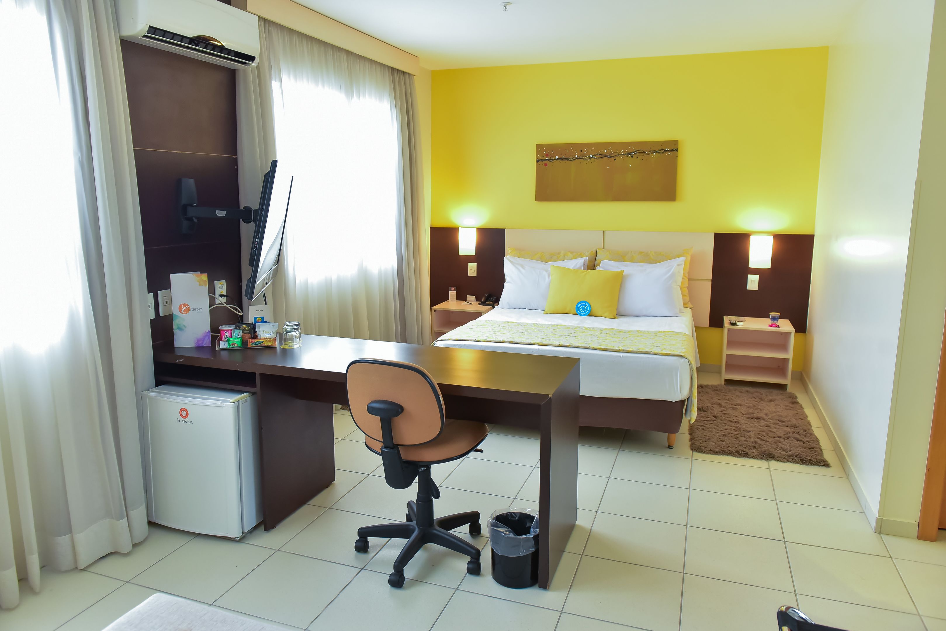 A hotel room with a bed, desk, chair, and a small refrigerator.