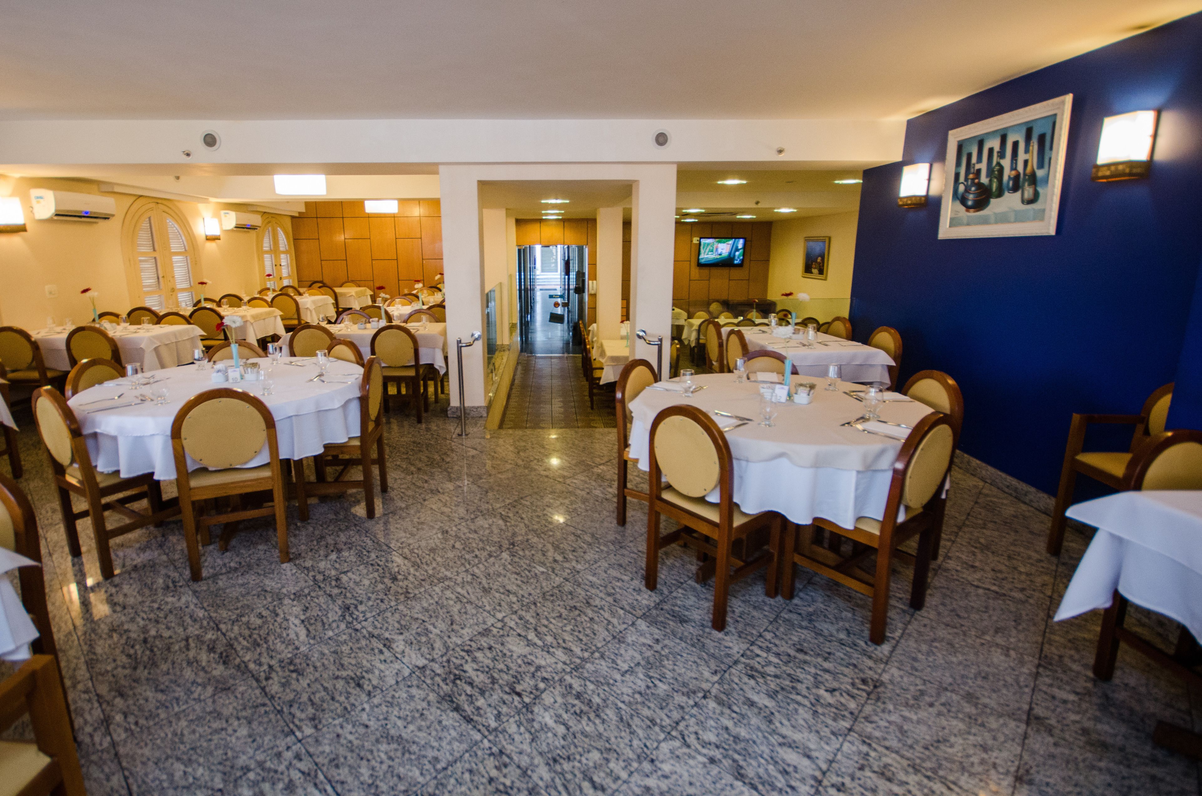 Un restaurante con varias mesas y sillas, piso de baldosas y una pared azul con un cuadro.