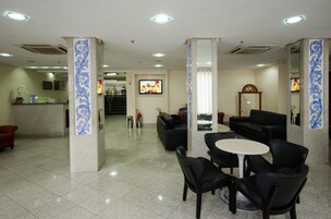 Un hall de hotel con un mostrador de recepción, sofás negros, una pequeña mesa blanca y un televisor montado en la pared.