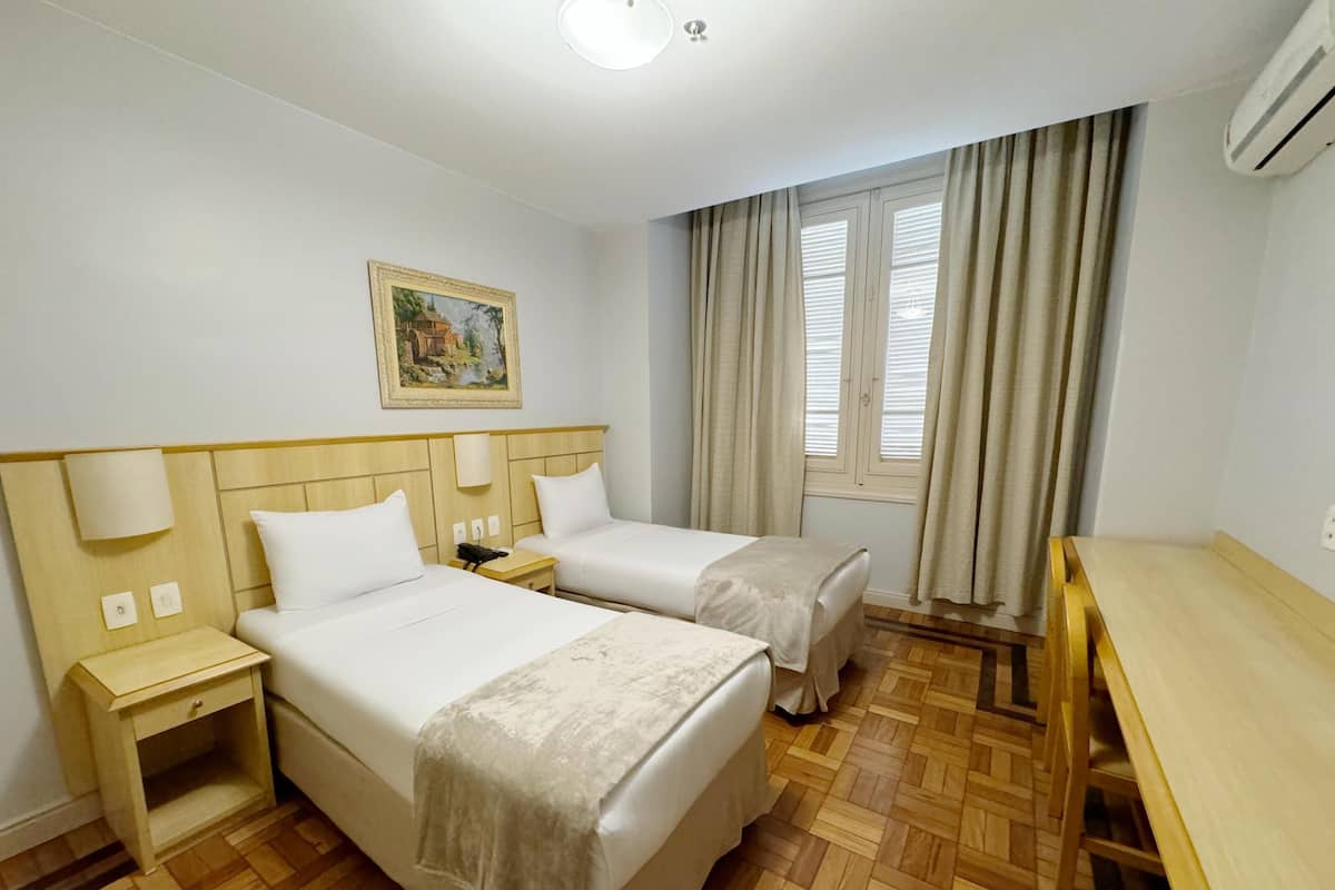Ein Hotelzimmer mit zwei Betten, einem Schreibtisch, einem Stuhl, einem Nachttisch, einer Lampe, einem Bild an der Wand und einem mit Vorhängen versehenen Fenster.
