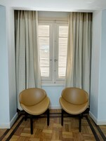 Quarto com duas cadeiras de couro marrom, uma janela com persianas brancas e chão de taco de madeira.