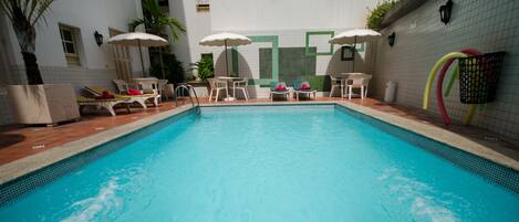 A swimming pool with lounge chairs and umbrellas, surrounded by a tiled wall and a small garden area.