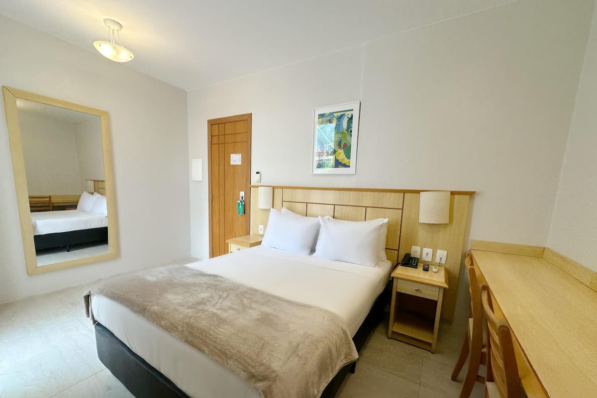 Ein Hotelzimmer mit einem großen Bett, einem Nachttisch mit Lampe, einem Schreibtisch, einem Spiegel und einer Tür.