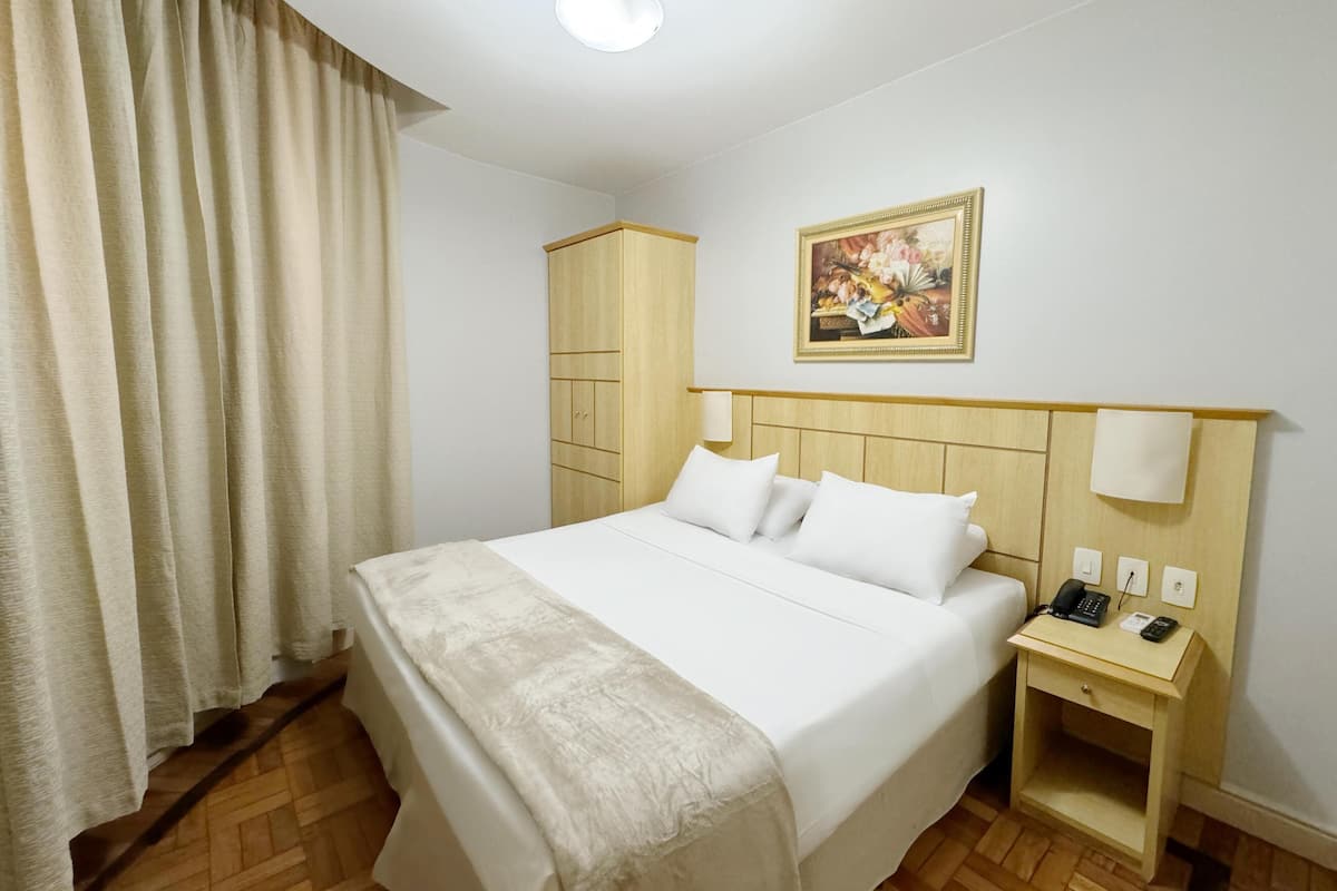 Ein Hotelzimmer mit Bett, Nachttisch, Kleiderschrank, einem Gemälde und einem Vorhang.