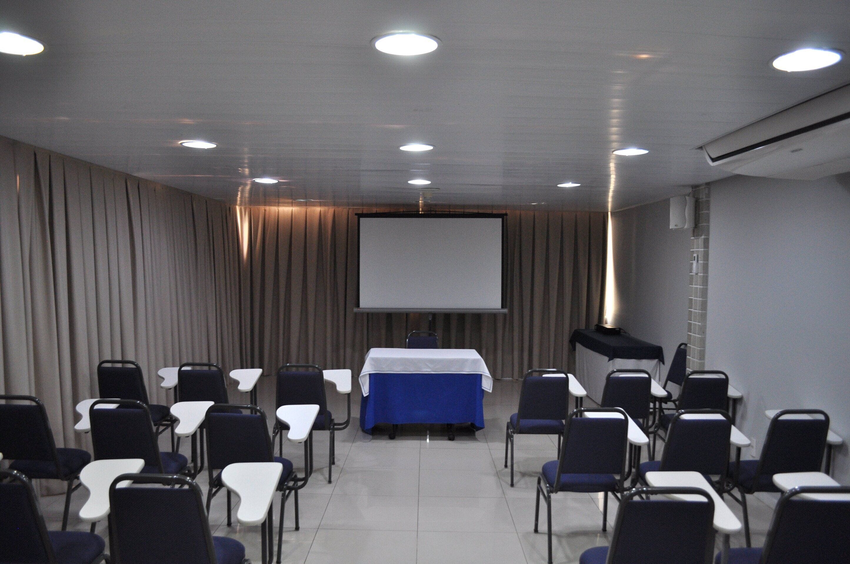 Un salón de conferencias con una pantalla de proyector, una mesa con mantel azul y filas de sillas.