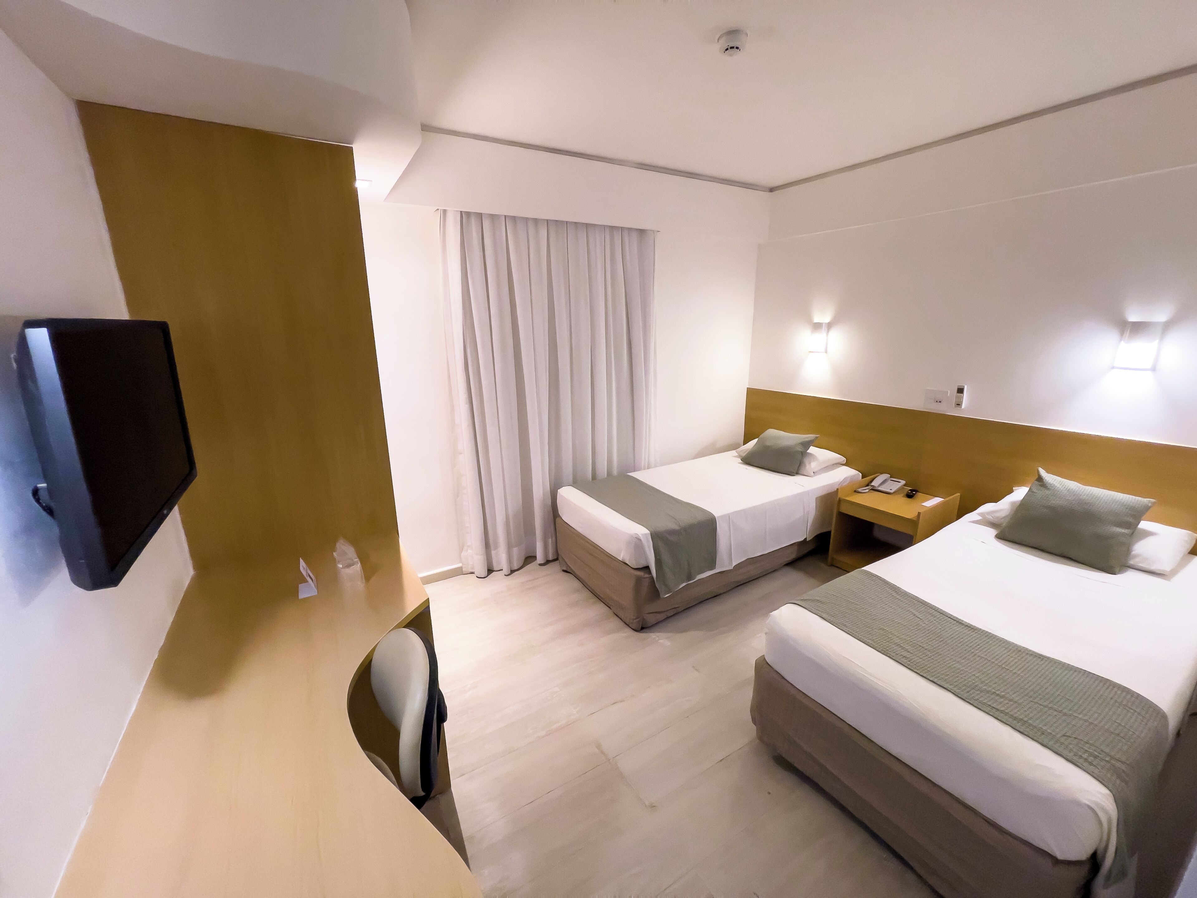Quarto de hotel com duas camas, televisão, escrivaninha e janela com cortinas.