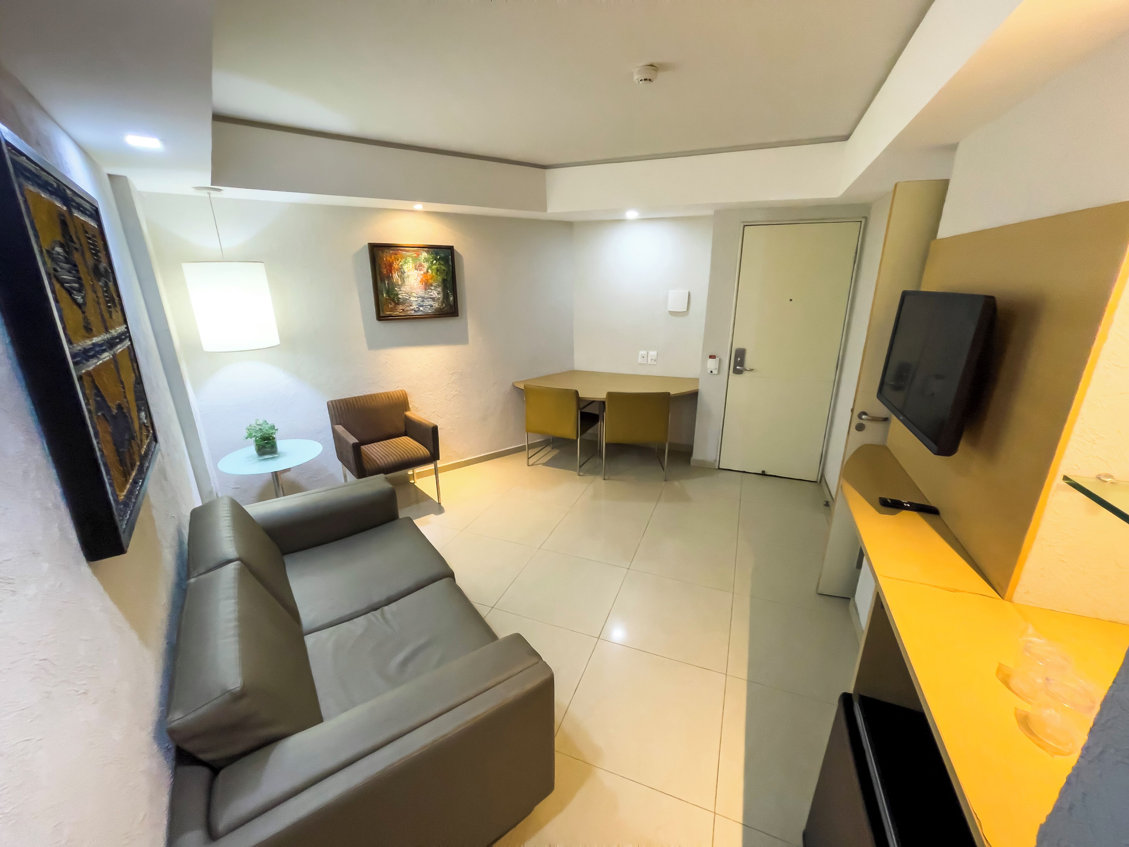 A modern living room with a sofa, a small table with a plant, a chair, a desk, and a television mounted on the wall.