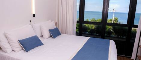 A bed with white and blue bedding, a bedside table, and a view of the ocean through a large window.