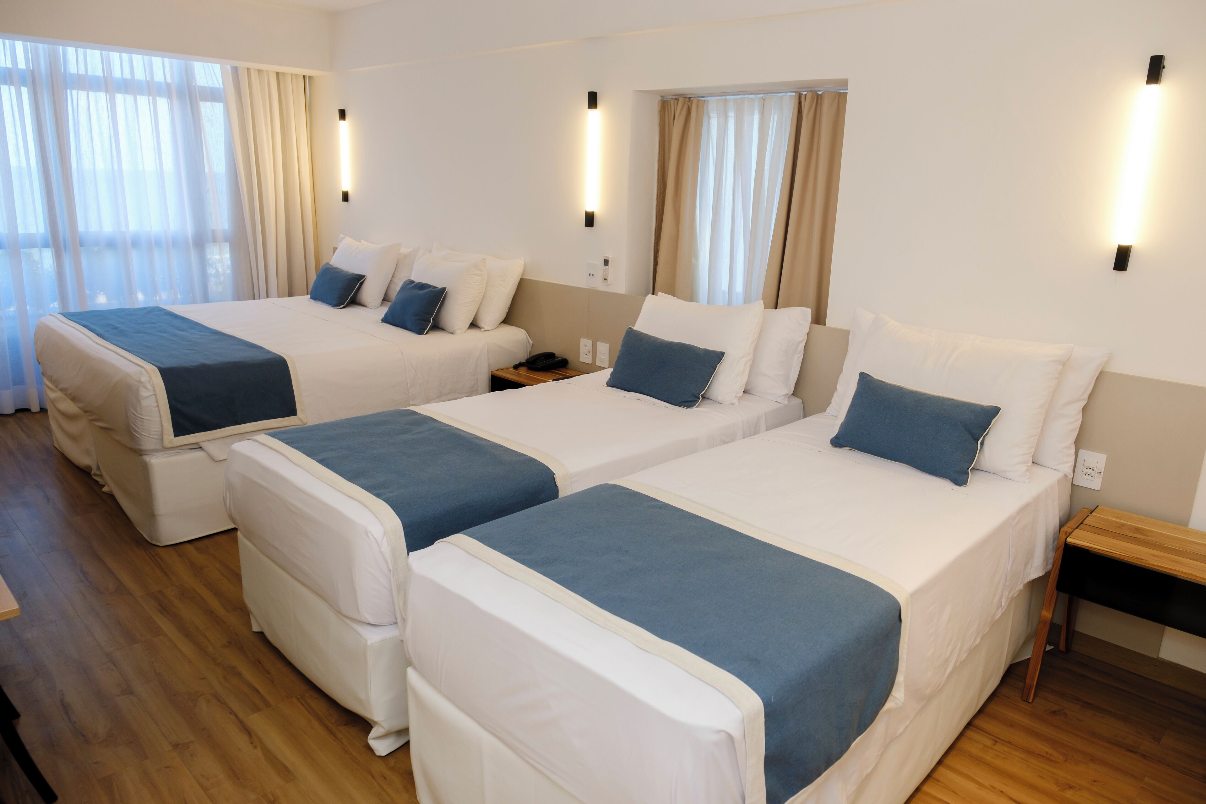 A hotel room with three single beds, each with a blue blanket and white bedding. There are bedside tables with lamps and a wooden nightstand.