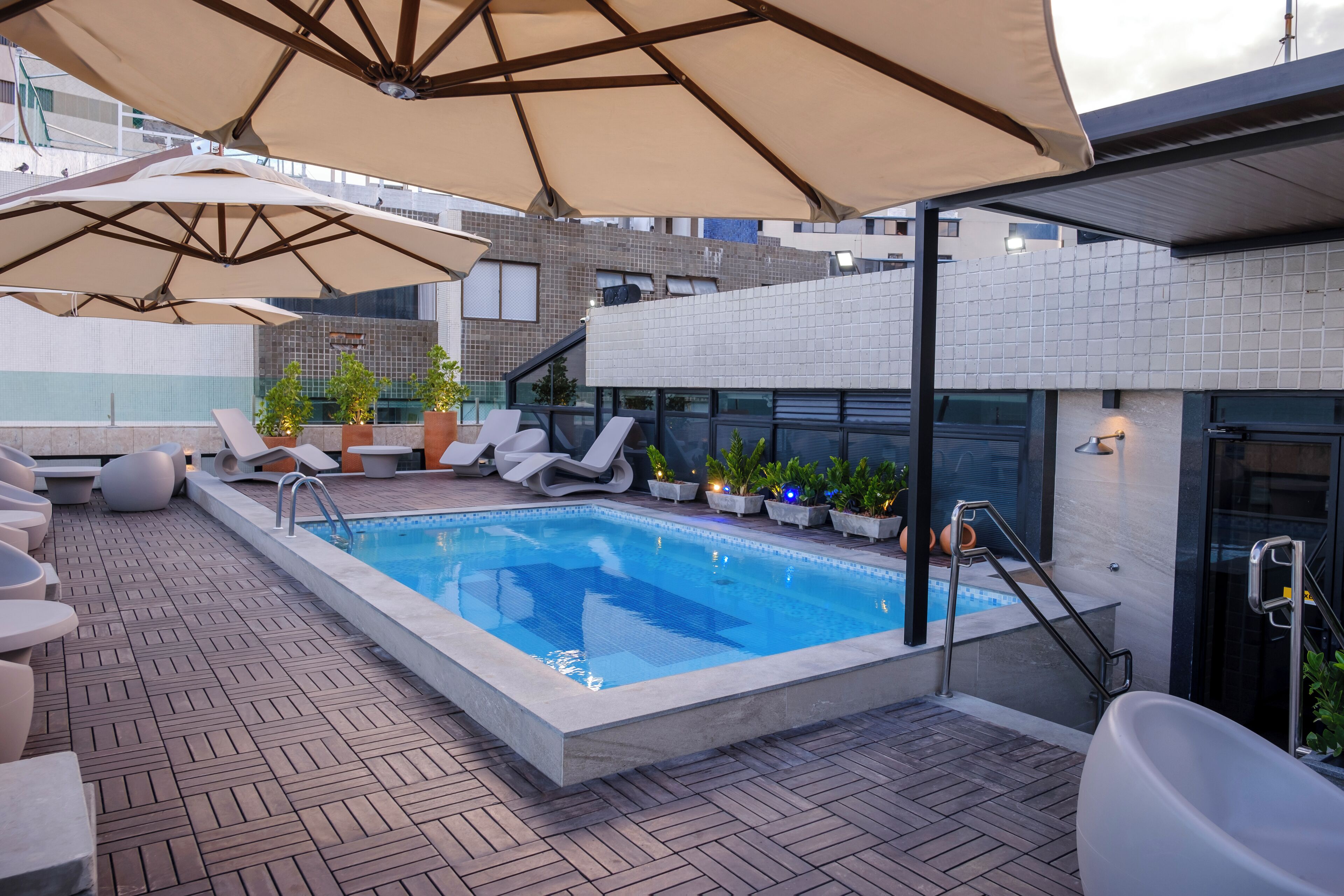 A rooftop pool area with lounge chairs, umbrellas, and potted plants.