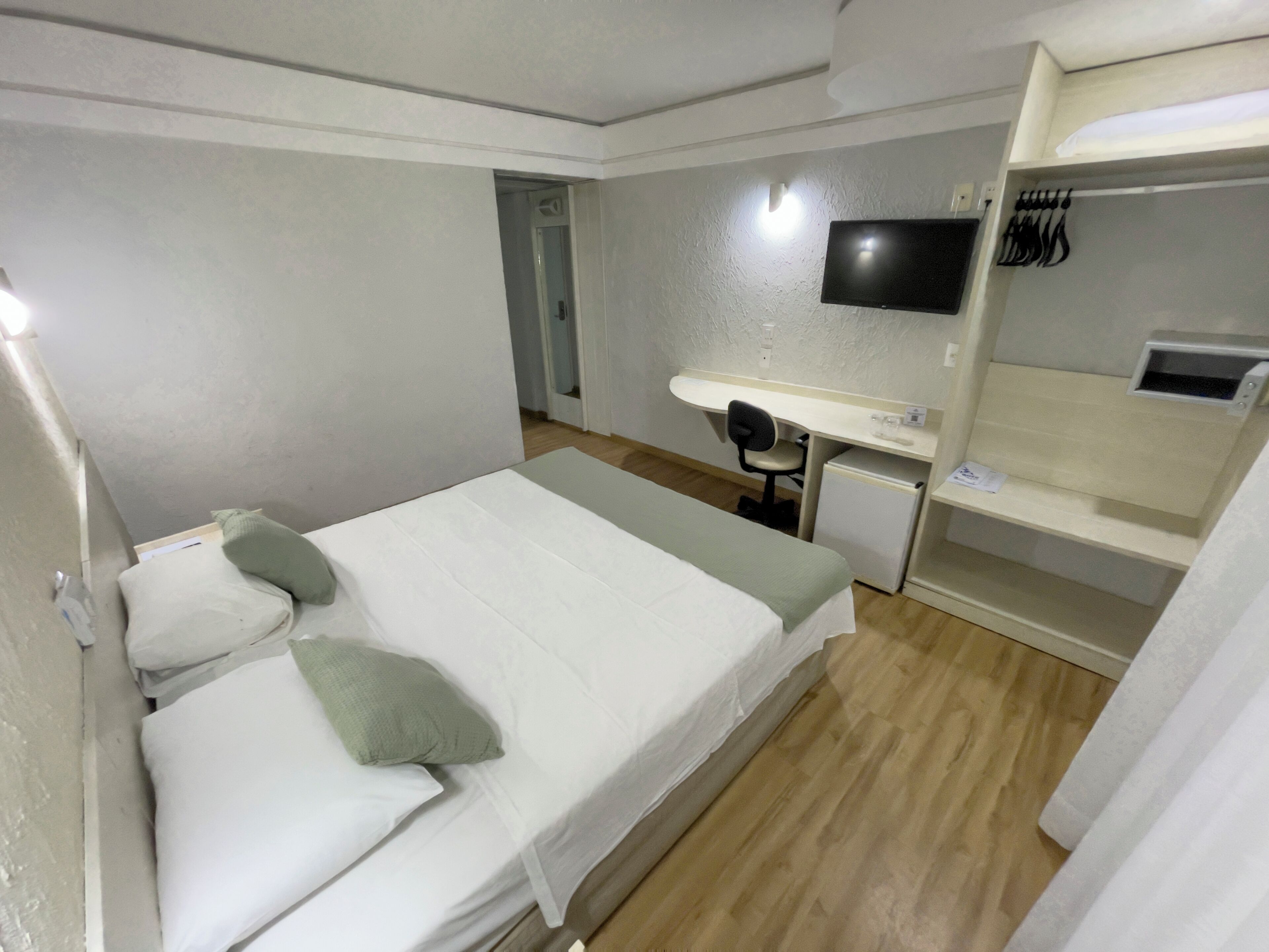 Quarto de hotel com uma cama, escrivaninha, cadeira, televisão e um pequeno frigorífico.