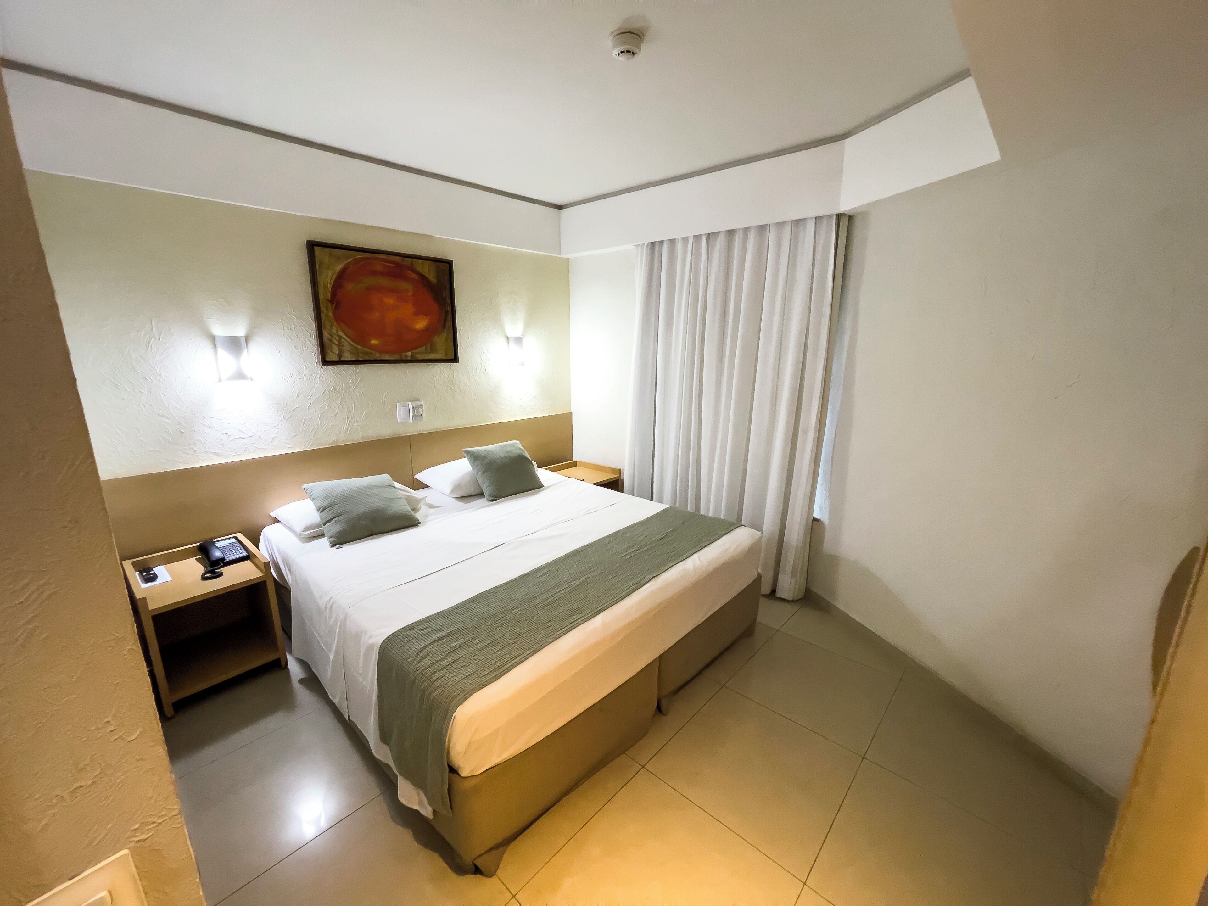 A hotel room with a large bed, a nightstand with a phone, a painting on the wall, and a window with curtains.