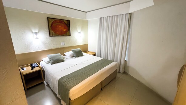 A hotel room with a large bed, a nightstand with a phone, a painting on the wall, and a window with curtains. - Marante Plaza Hotel (Recife)
