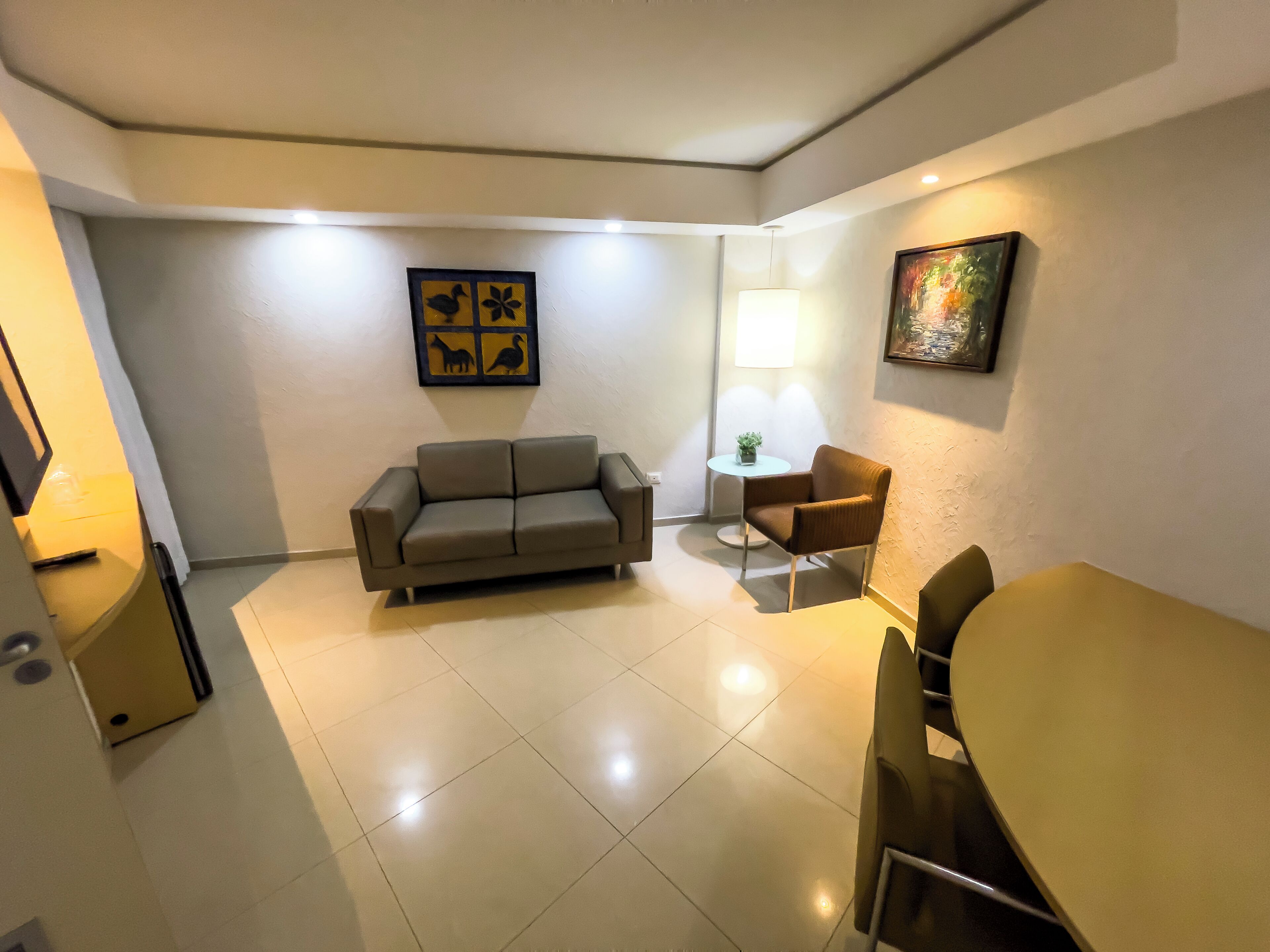A compact living area with a sofa, a small round table, and a chair. There is a lamp and a framed artwork on the wall.