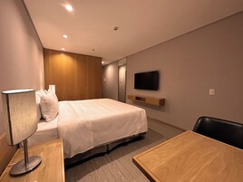 Una habitación de hotel con cama, escritorio, silla, televisor montado en la pared y armario de madera.