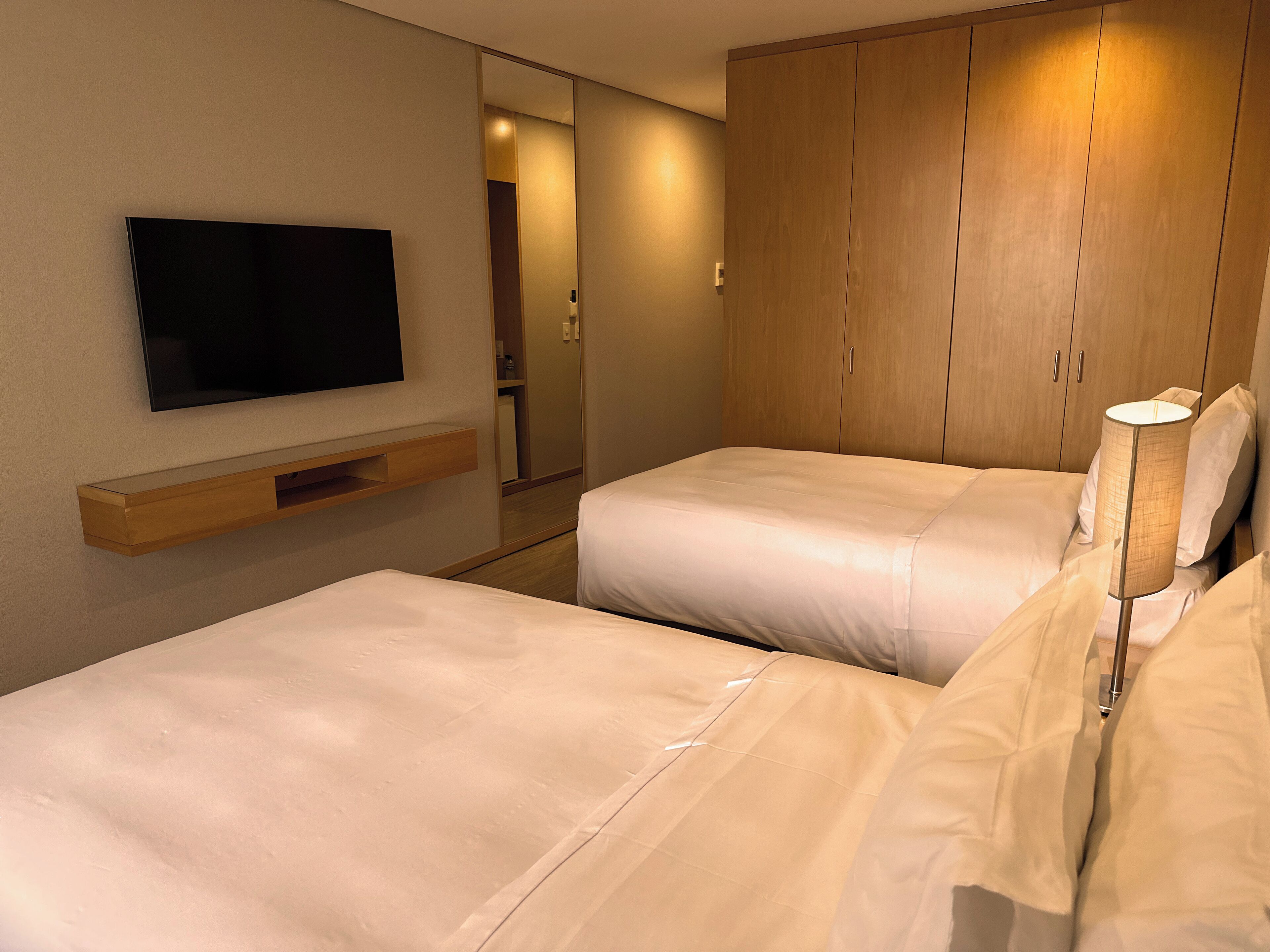 Ein Hotelzimmer mit zwei Betten, einem an der Wand montierten Fernseher, Holzschränken und einer Nachttischlampe.
