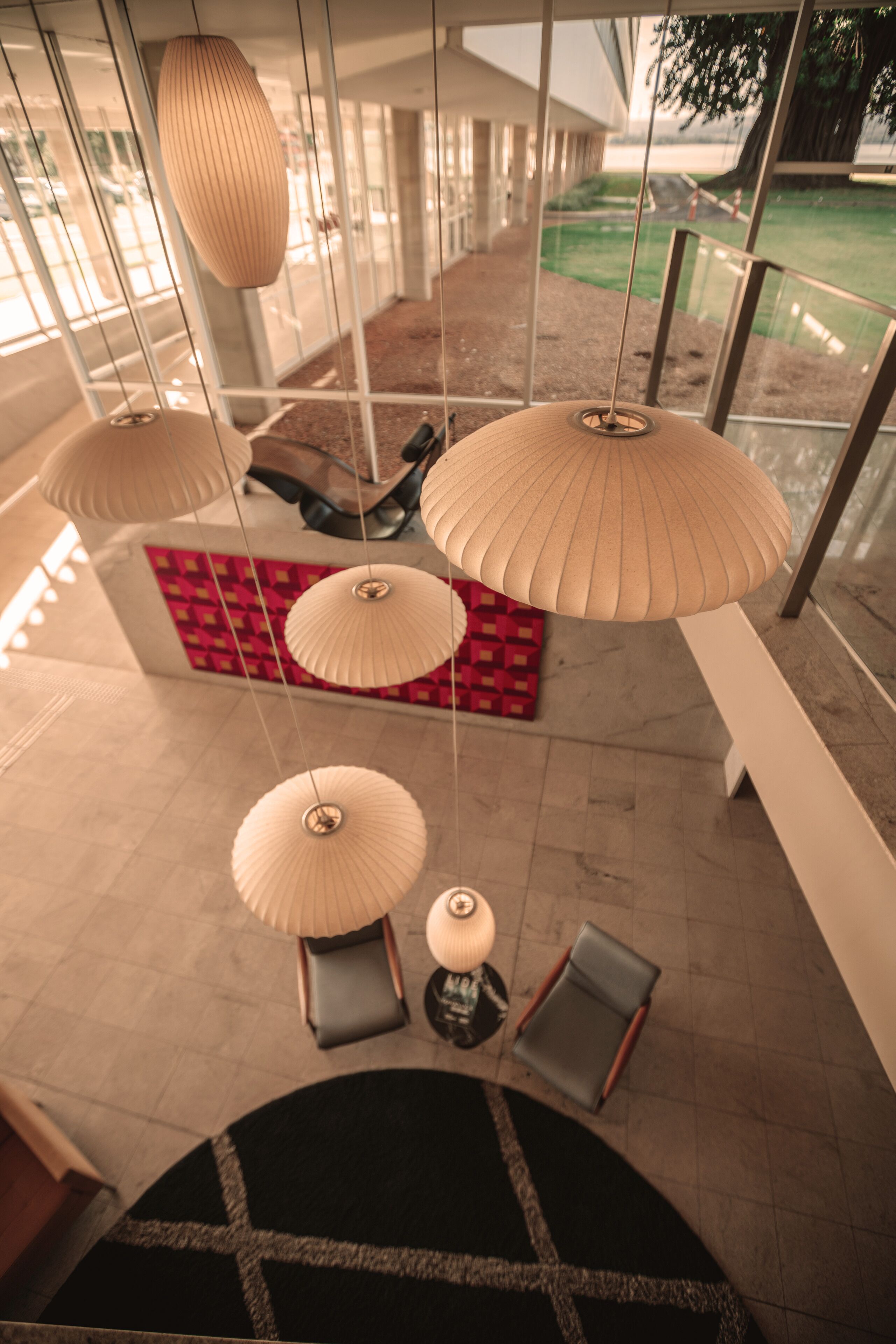 A modern interior with hanging paper lamps, a red patterned rug, and a view of a green outdoor area.