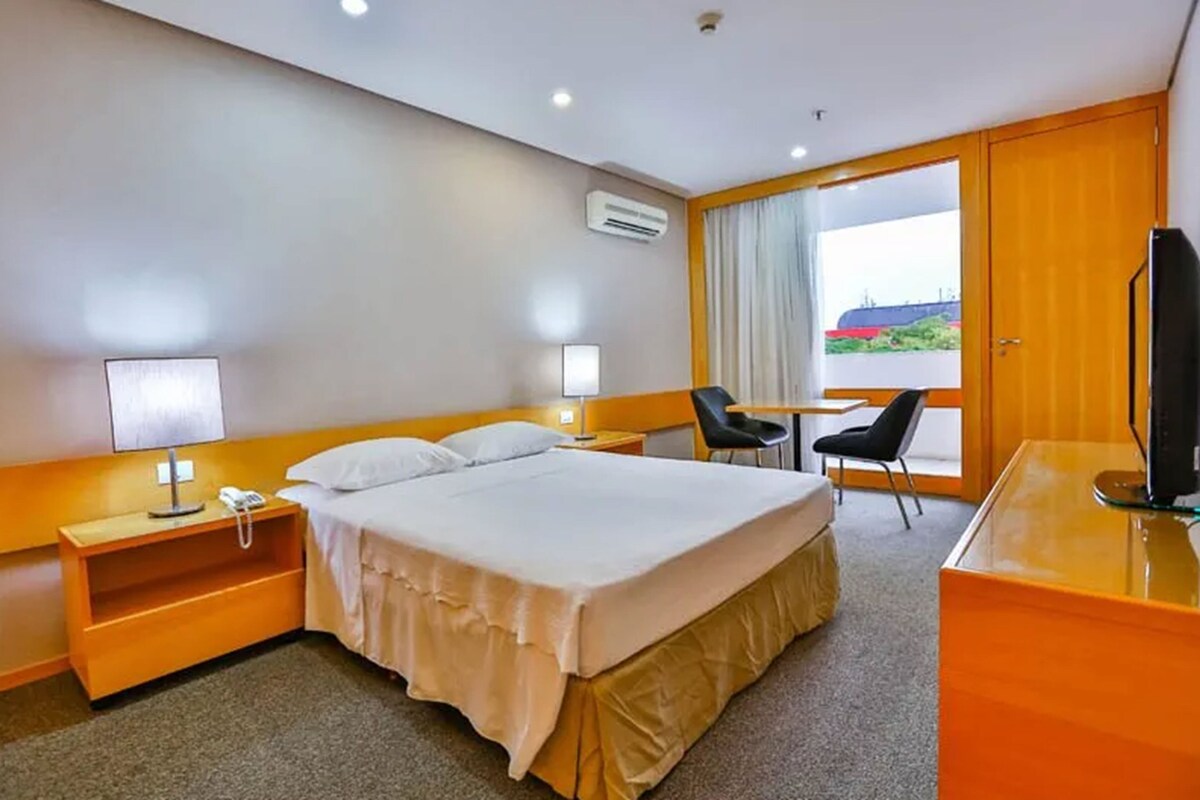 A hotel room with a bed, bedside tables, a desk, a chair, a TV, and a large window with a view.