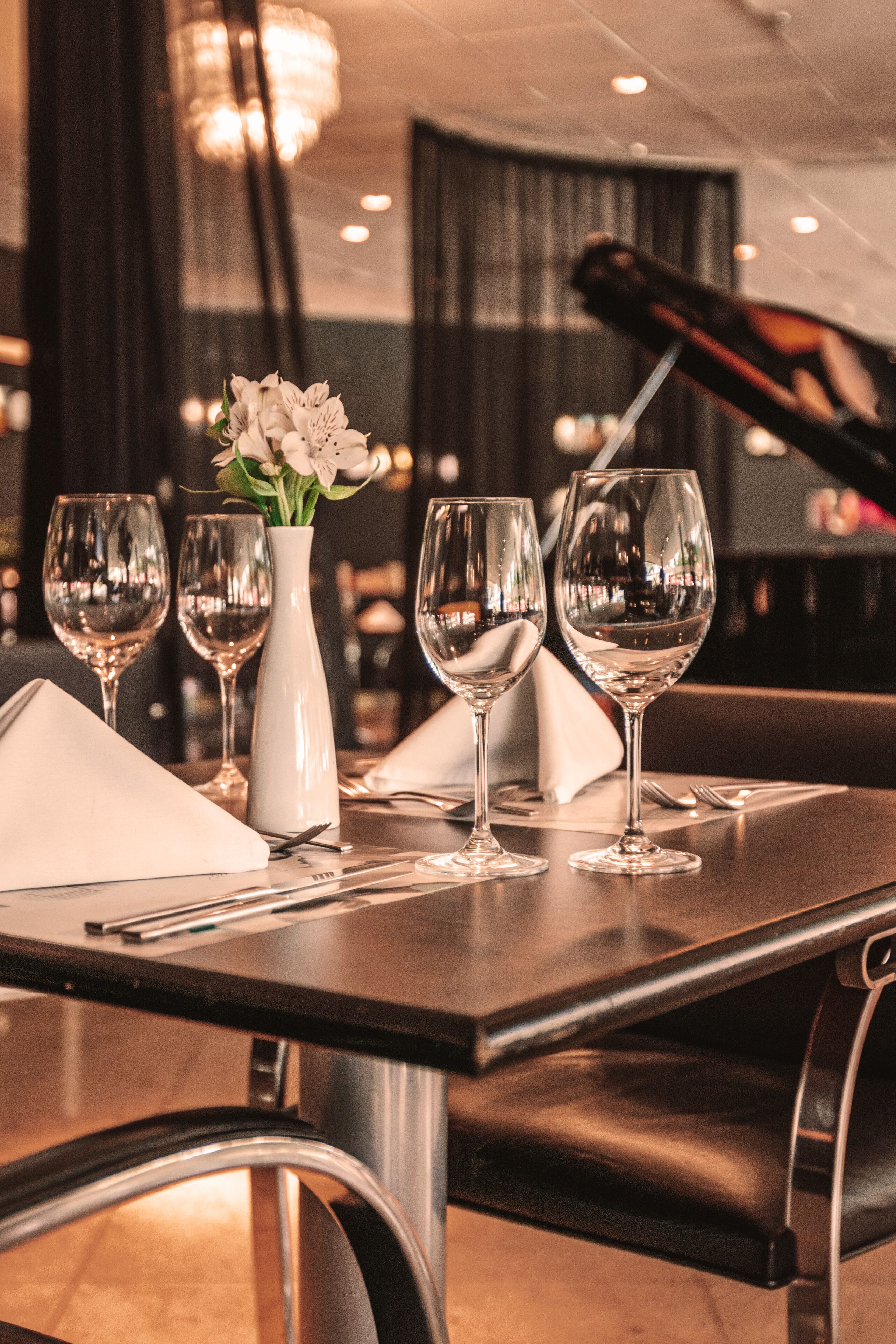 Una mesa de comedor dispuesta con copas de vino y un florero, con un piano al fondo.