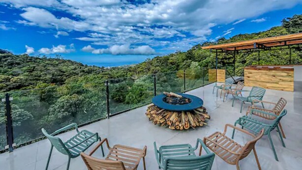 A rooftop terrace with a fire pit, outdoor seating, and a glass balustrade overlooking a lush landscape.