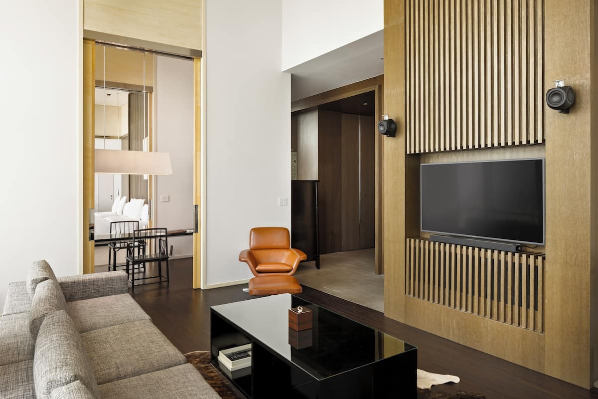 A modern living room with a sectional sofa, a glass coffee table, a leather armchair, a flat-screen TV mounted on a wooden panel, and a dining area with a lamp and chairs.