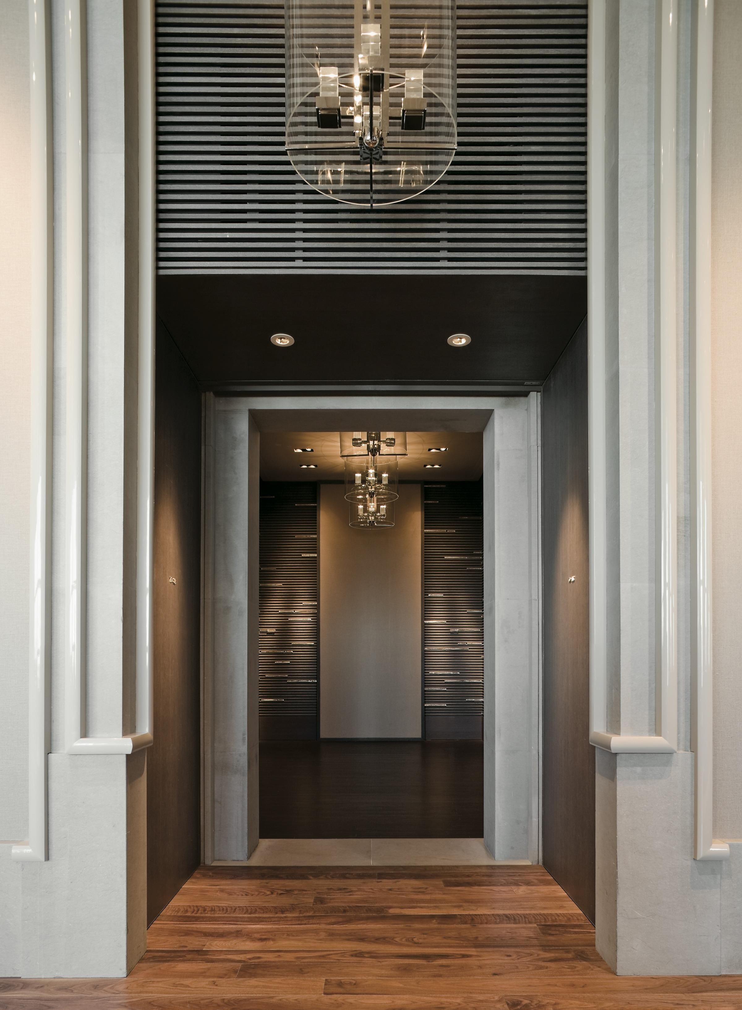 Um corredor com um lustre moderno, piso de madeira e vista para um quarto com parede escura e uma porta.
