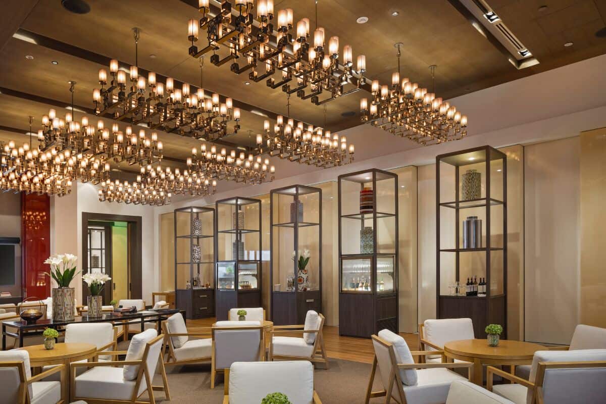 A spacious lounge area with multiple seating arrangements, including sofas and tables. There are decorative light fixtures hanging from the ceiling, and built-in shelving units with various items displayed.