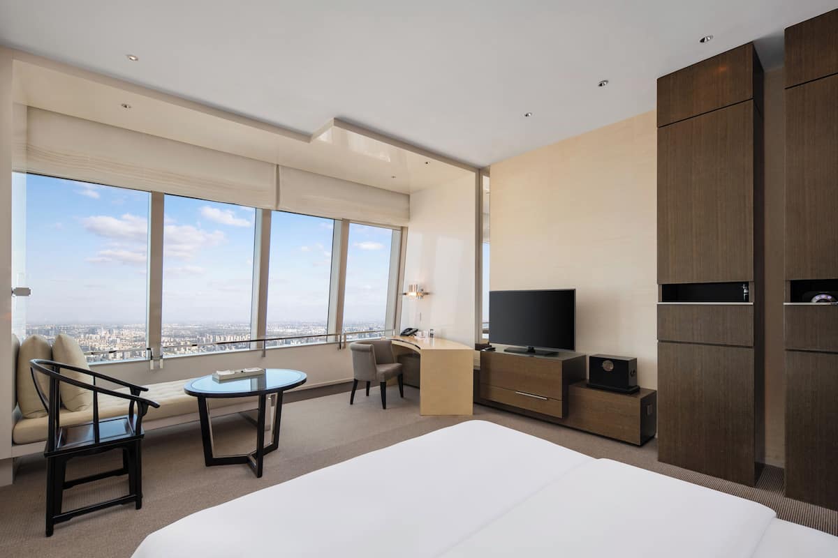 A modern hotel room with a large bed, a desk, a chair, a TV, and a view of the city.