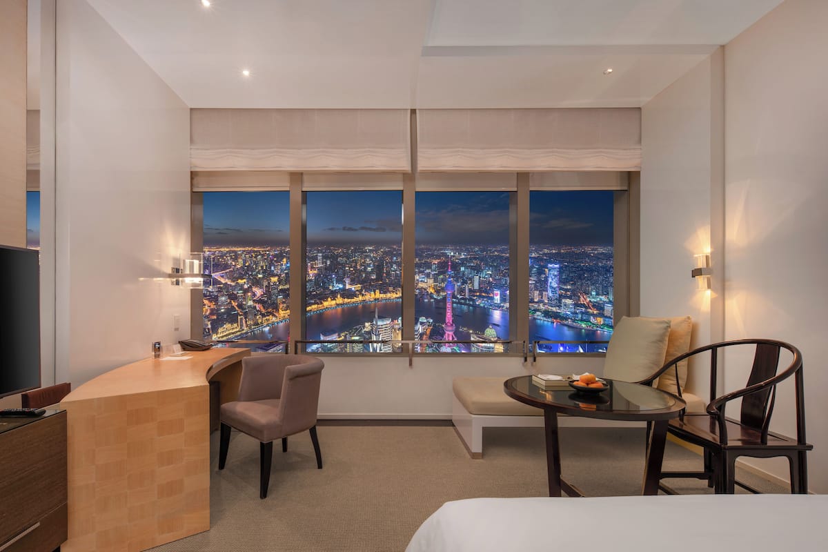 A hotel room with a large window offering a city view, a desk, a chair, a small table, and a bed.