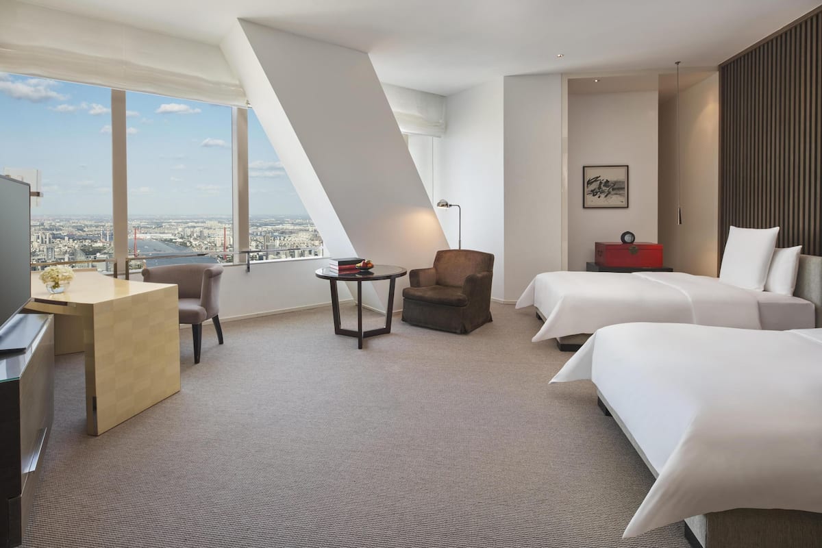 A hotel room with a desk, two beds, a chair, a small table, and a view of the city.
