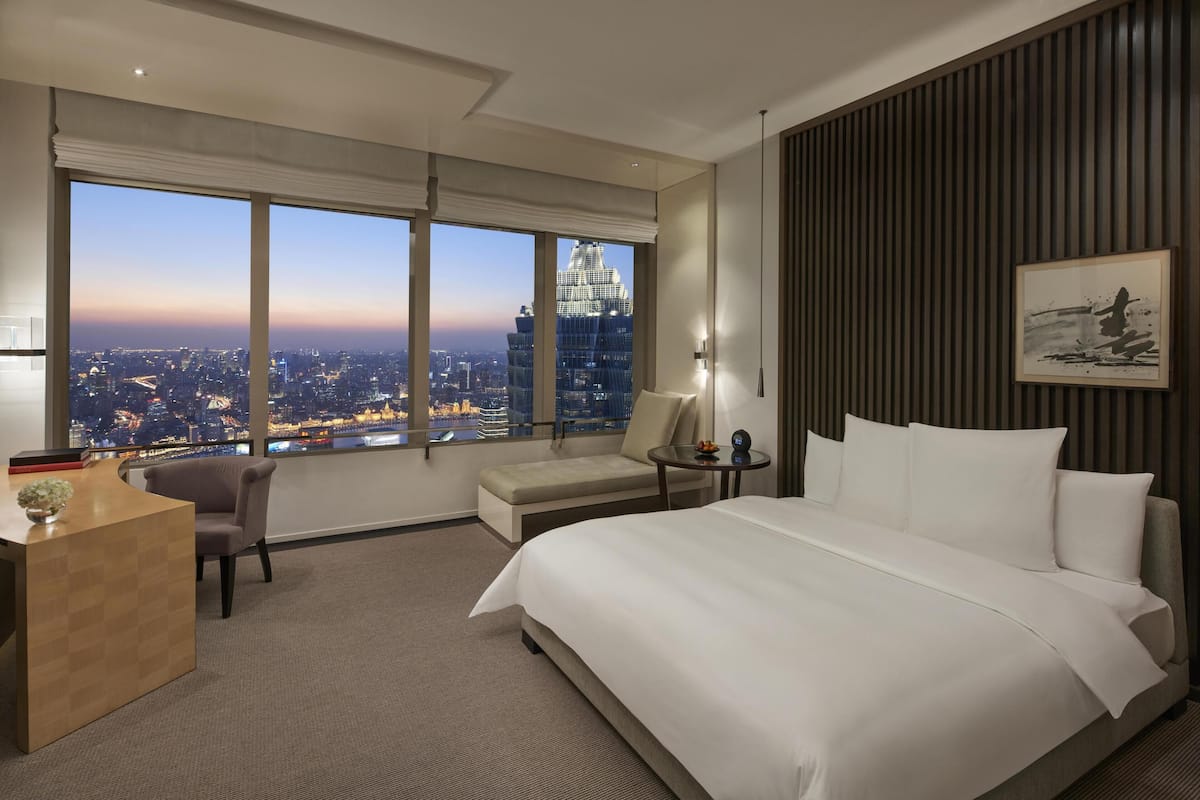 A hotel room with a large bed, a desk, a chair, a small table, and a large window offering a city view.