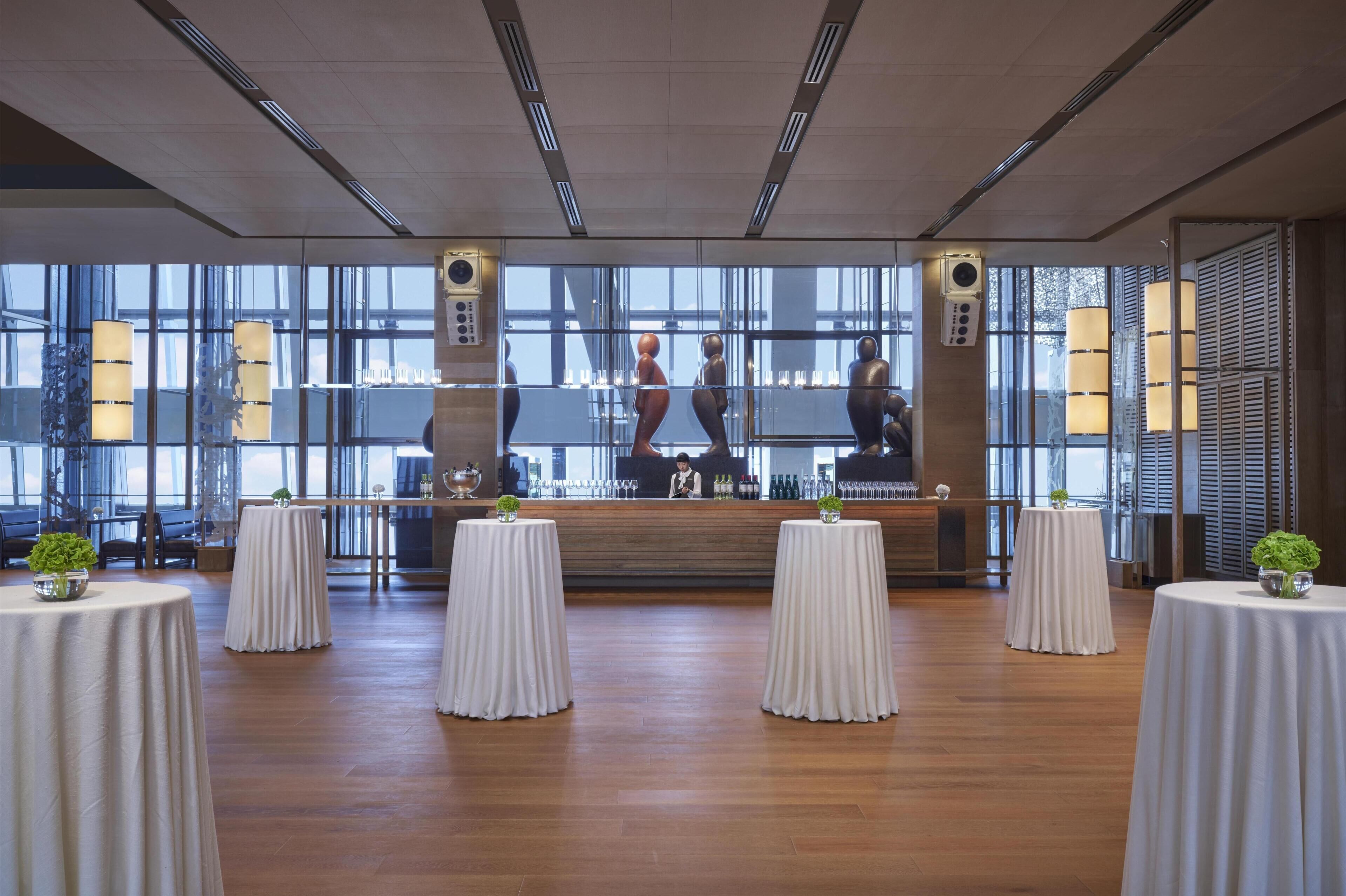 Une pièce spacieuse avec de grandes fenêtres, une scène avec un barman et plusieurs tables rondes recouvertes de nappes blanches.