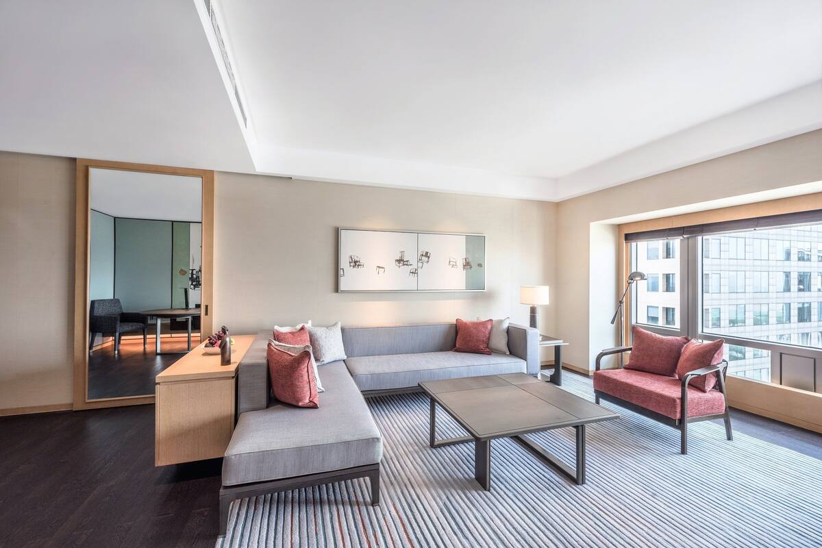 Une chambre d’hôtel moderne avec un canapé gris, une table basse, une petite table d’appoint et un grand miroir.