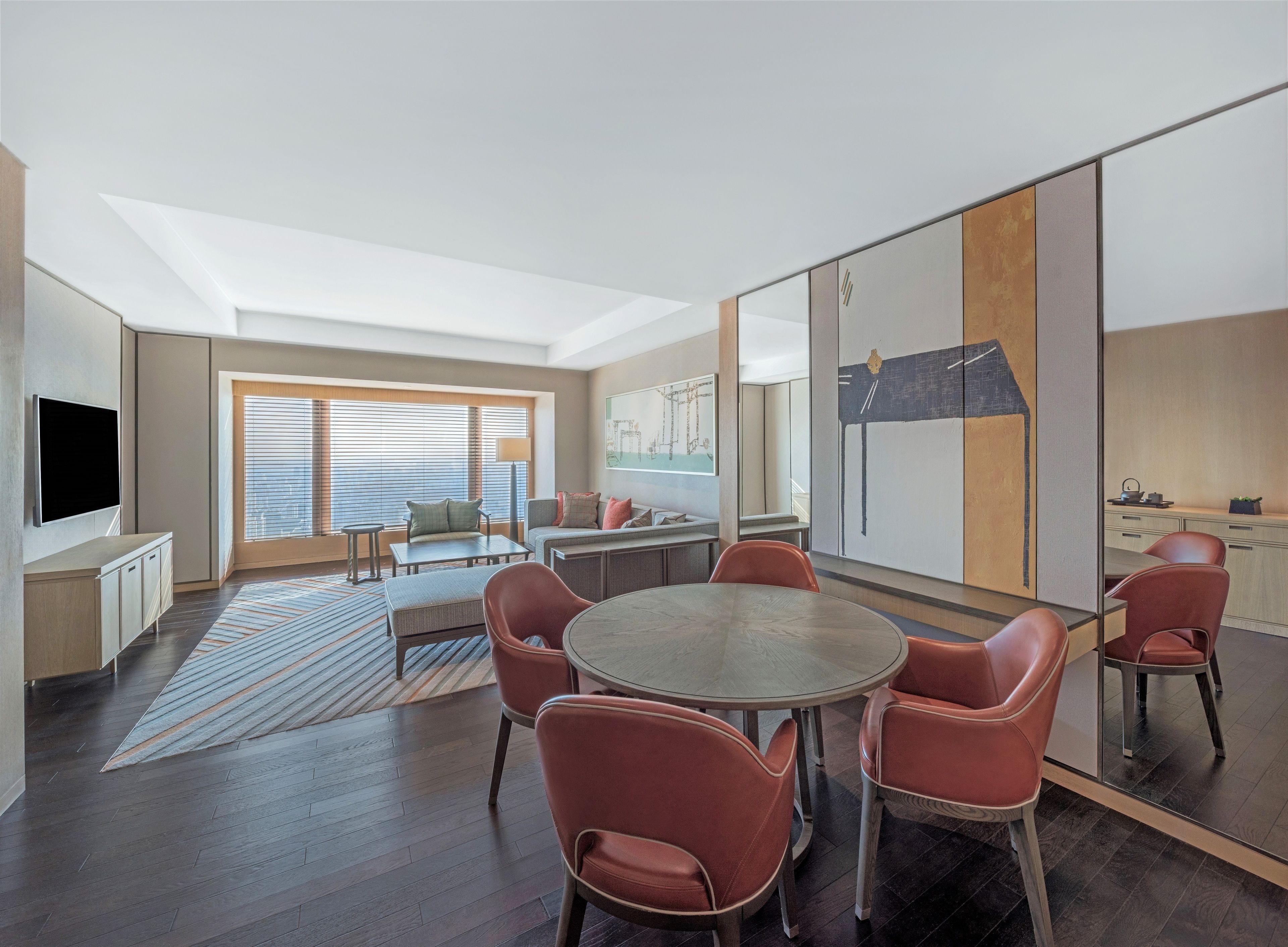A modern hotel room with a dining area, a sofa set, a TV, and a large window with blinds.