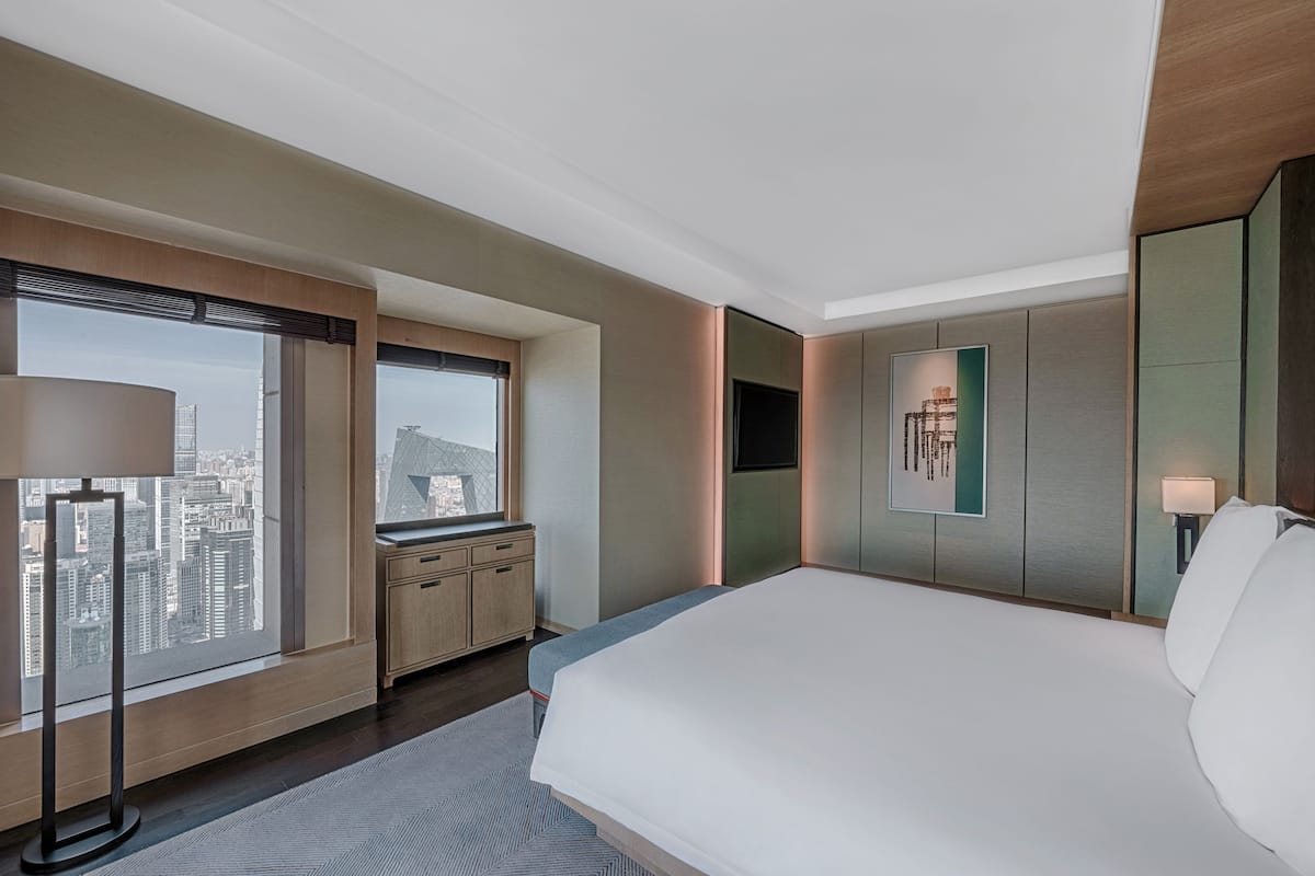 Une chambre d’hôtel moderne dotée d’un grand lit, d’un bureau, d’une chaise, d’une fenêtre donnant sur la ville et d’un téléviseur à écran plat fixé au mur.
