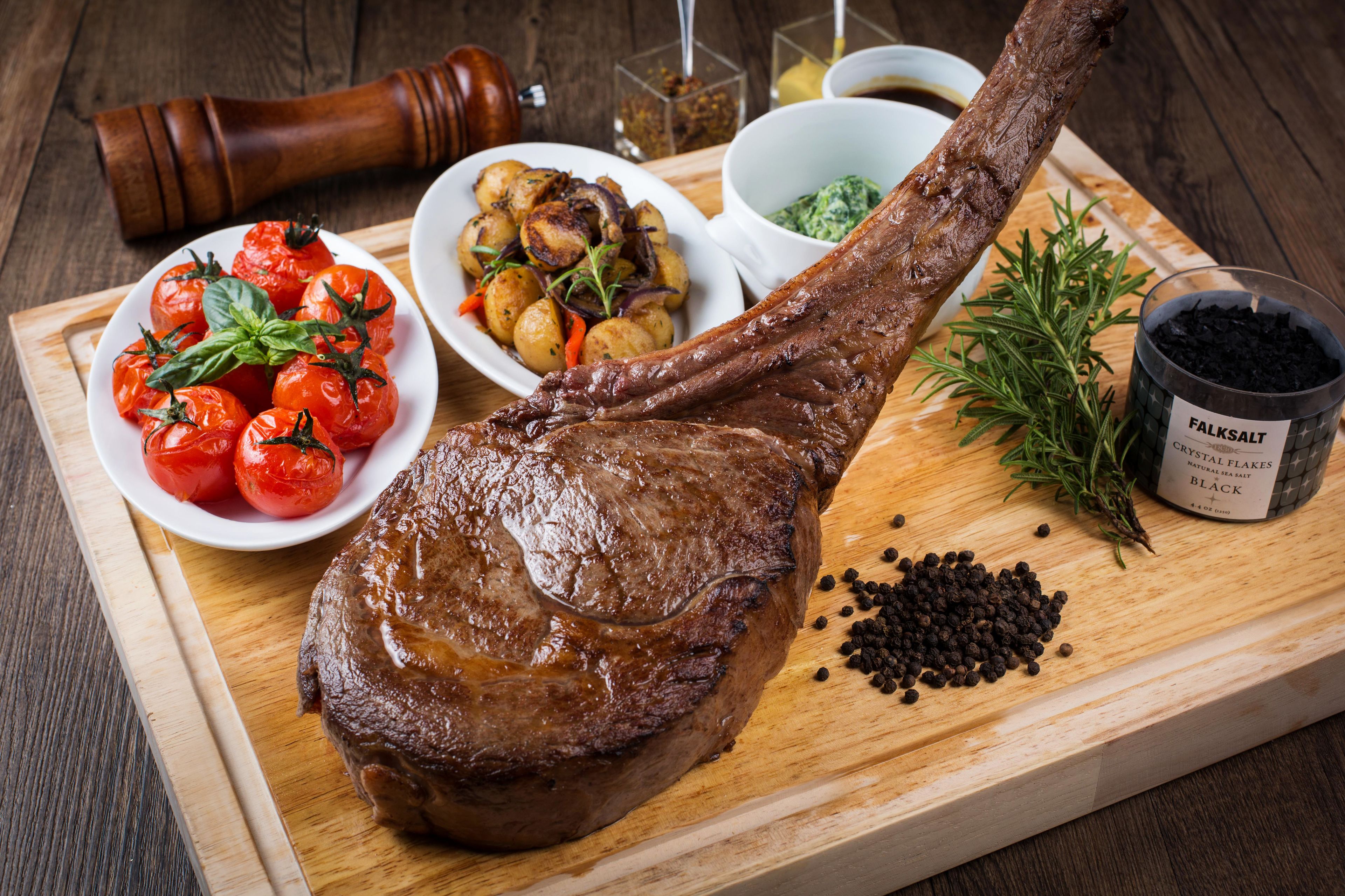 A wooden cutting board with a cooked lamb shank, roasted tomatoes, and a bowl of potatoes.