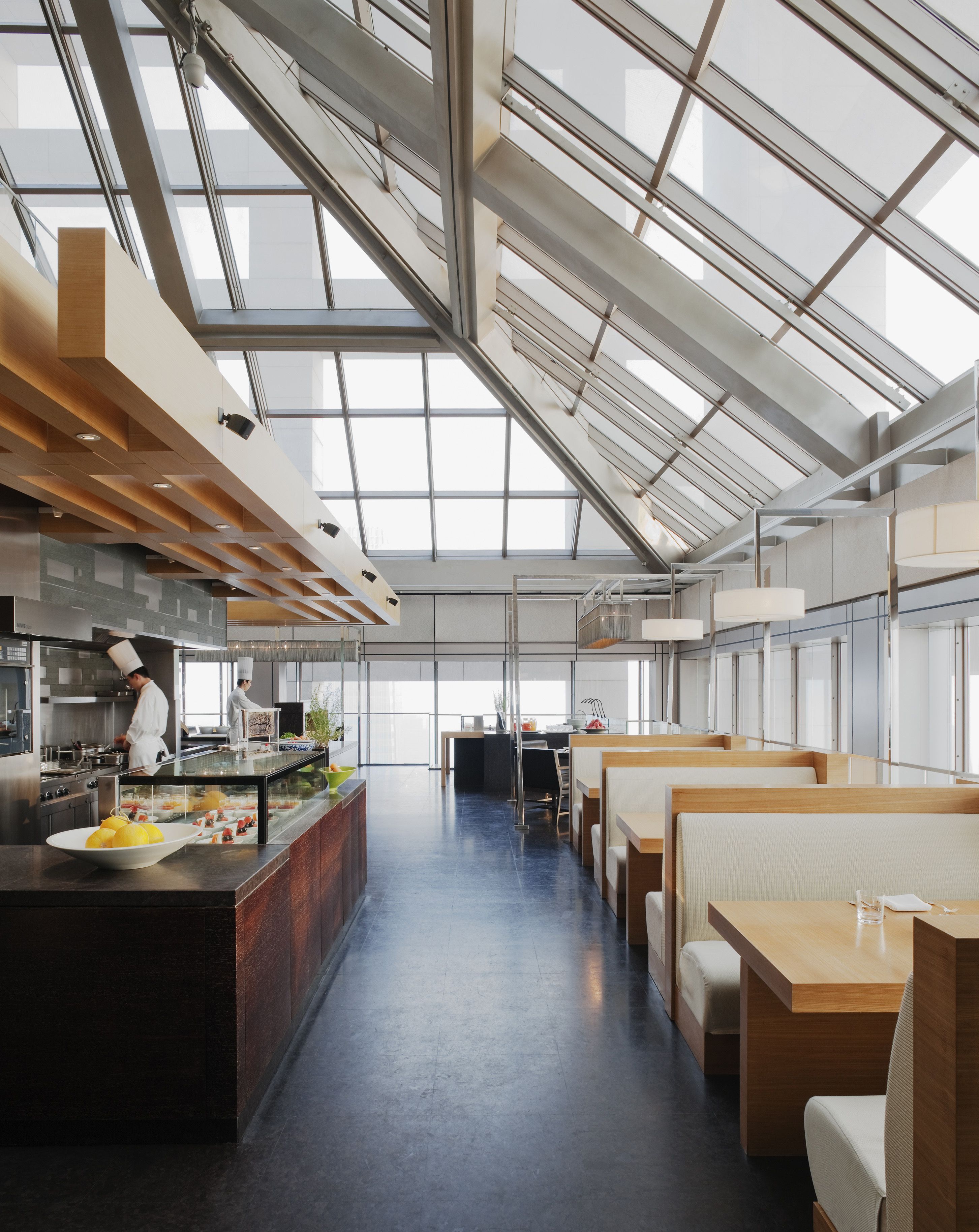 A modern restaurant with a high ceiling and large windows, featuring a central island with a chef preparing food.