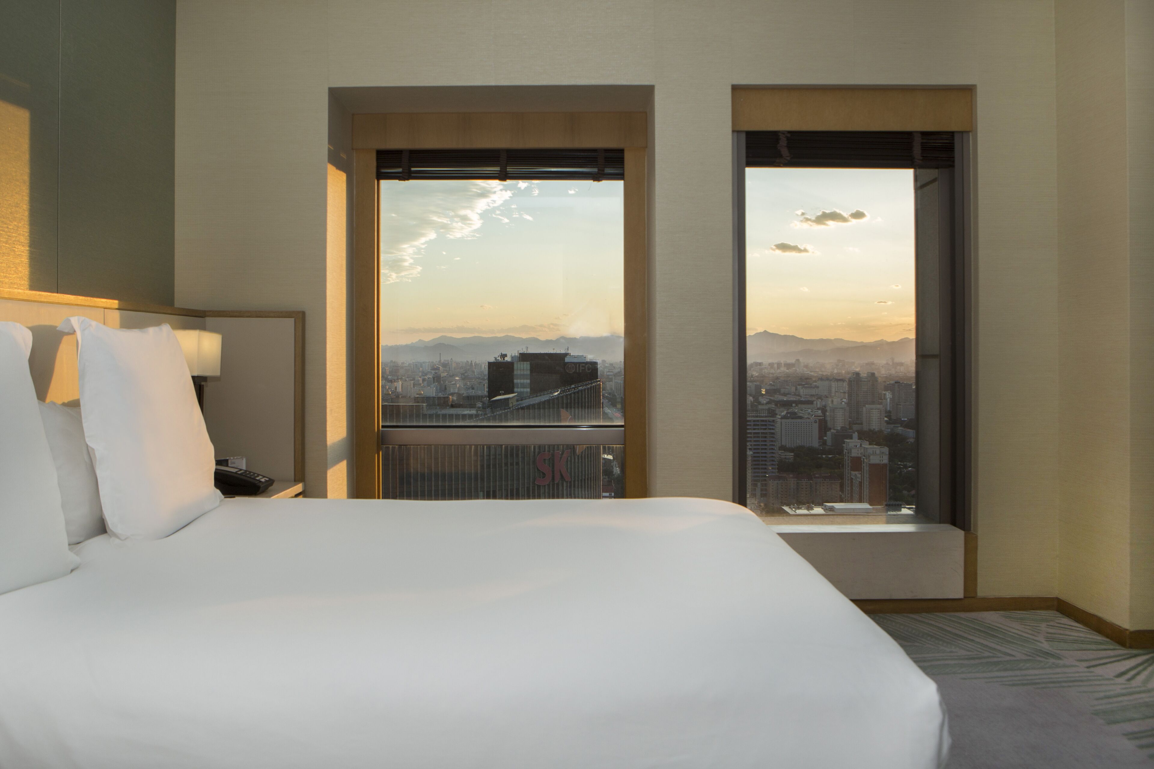 Une chambre d’hôtel avec un grand lit, une table de chevet et une vue sur le paysage urbain, visible à travers deux fenêtres.