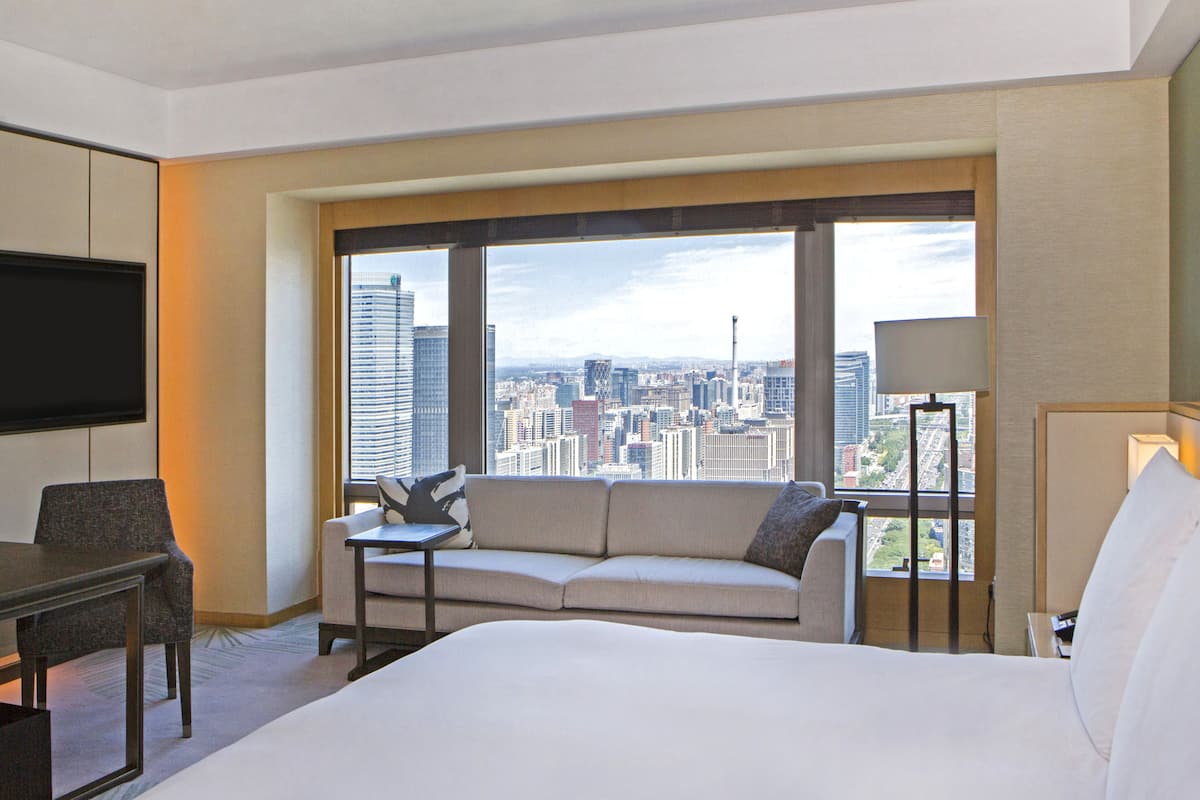 Une chambre d’hôtel avec un grand lit, un canapé, un bureau et une télévision. La chambre offre une vue sur la ville grâce à de grandes fenêtres.