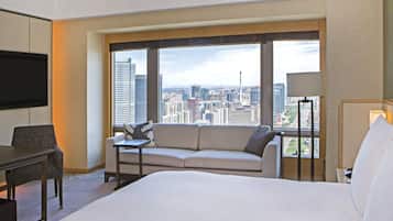 A hotel room with a large bed, a sofa, a desk, and a TV. The room has a city view through large windows.