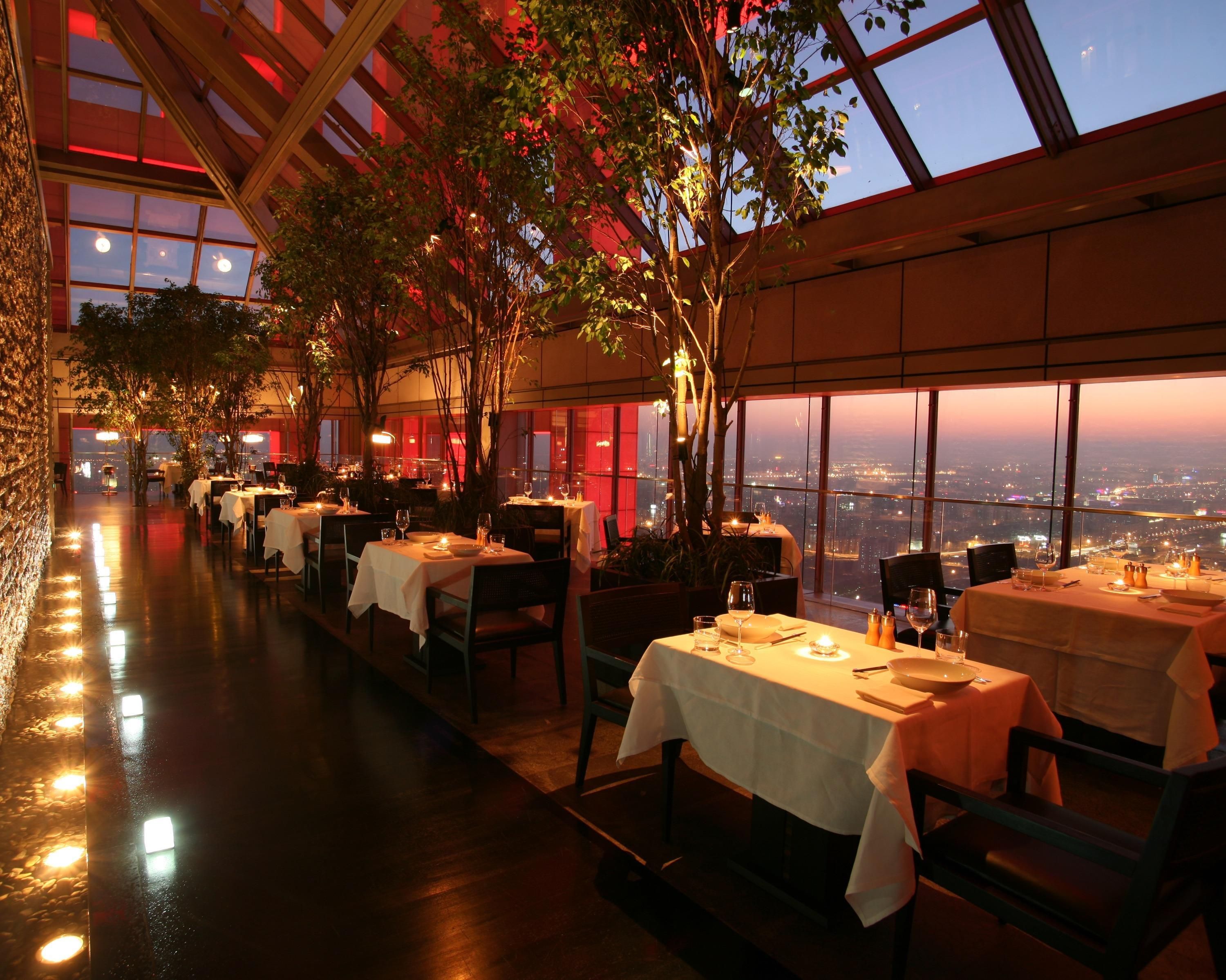 Un área de comedor interior con mesas dispuestas para los comensales, ventanas amplias que ofrecen vistas a la ciudad y una estructura de techo de madera.