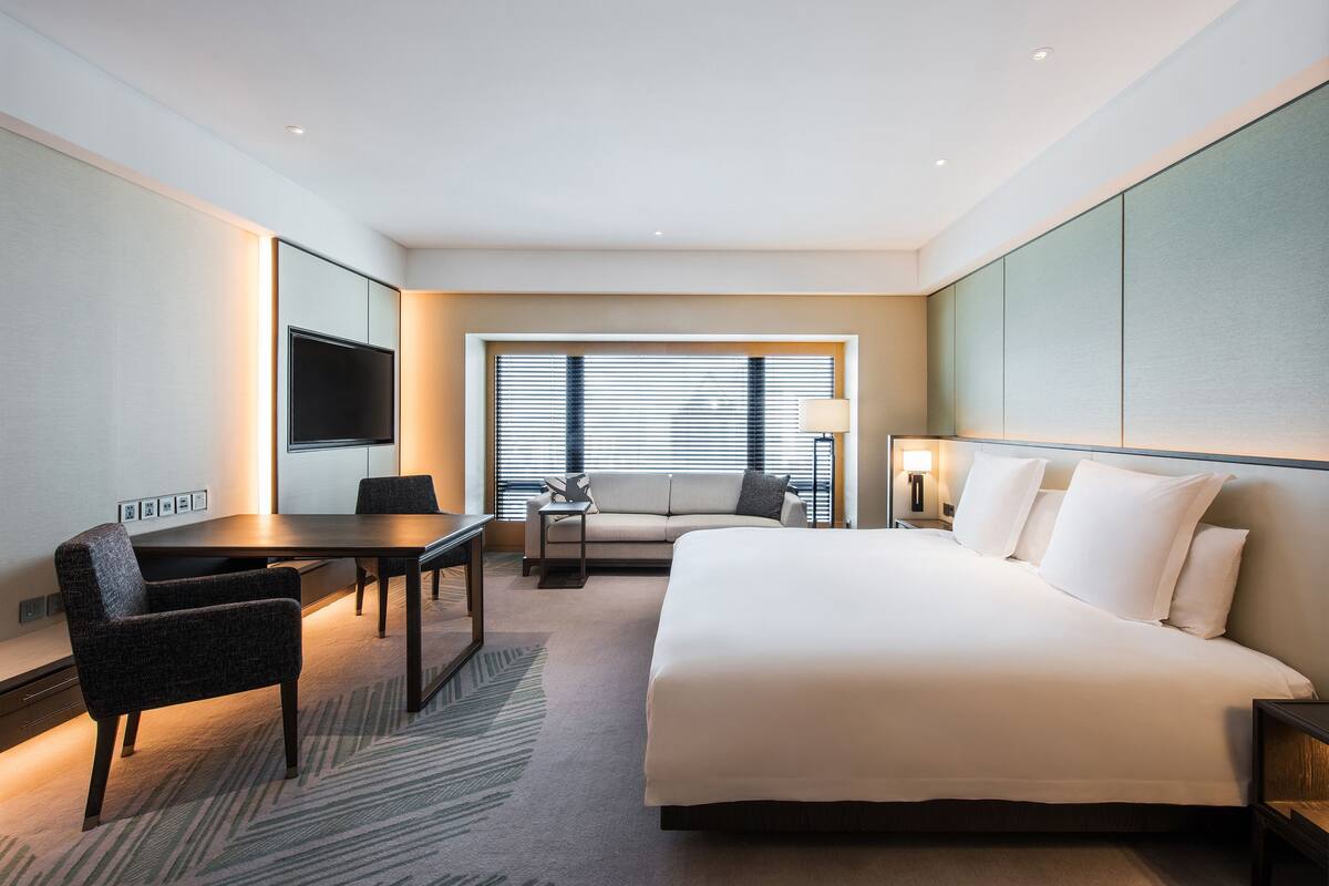 Une chambre d’hôtel avec un grand lit, un bureau, une chaise, une télévision et une fenêtre avec des stores.