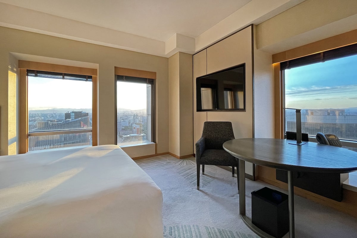 Une chambre d’hôtel équipée d’un lit, d’un bureau, d’une chaise, d’une télévision et de grandes fenêtres donnant sur la ville.
