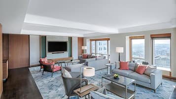 A modern hotel room with a large sofa set, a flat-screen TV mounted on the wall, and large windows offering city views.